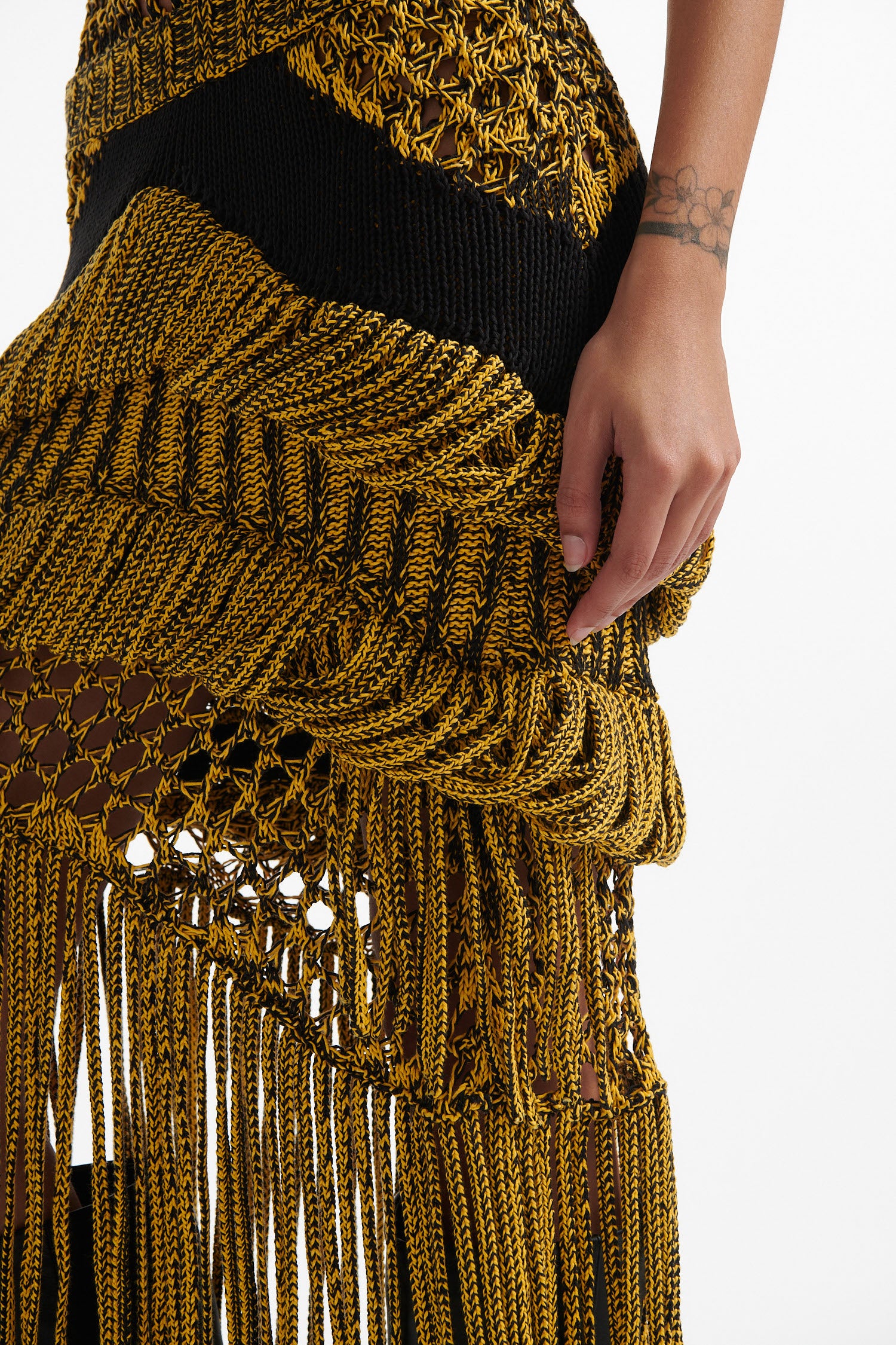 Female model wears yellow mesh tank and matching mixed-stitch skirt with black high heel boots on white background. 