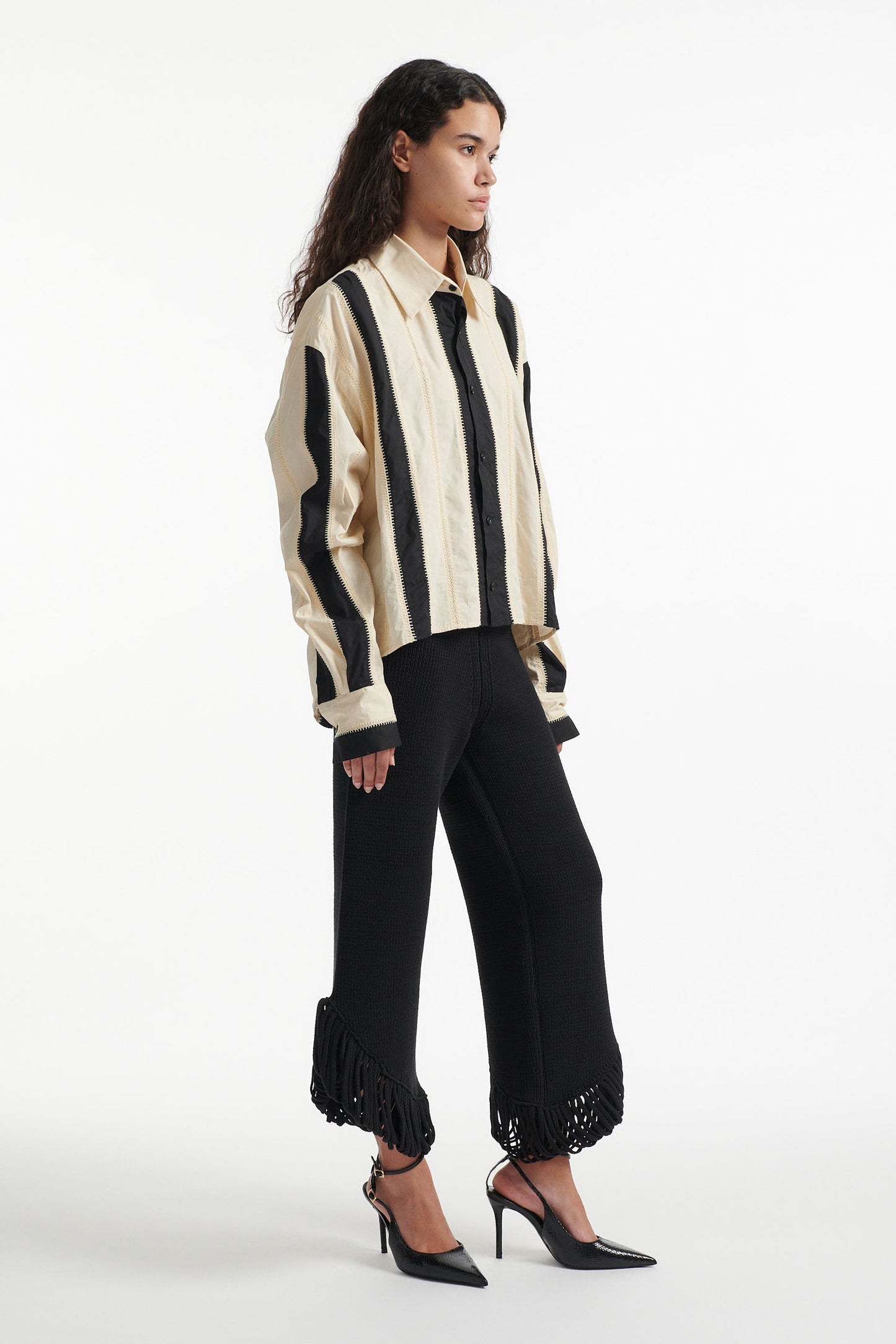 Female model wears off-white and black vertical striped panel button down shirt with cotton black pants and black high heel boots on white background. 