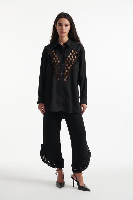 Female model wears black cotton shirt with see-through cutouts at chest and back. Model also wears black knit pants with fringe and black high heels on white background.