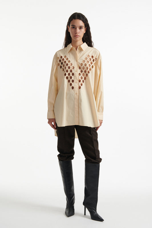 Female model wears off-white cotton shirt with see-through cutouts at chest and back. Model also wears brown wool pants and black high heels on white background.