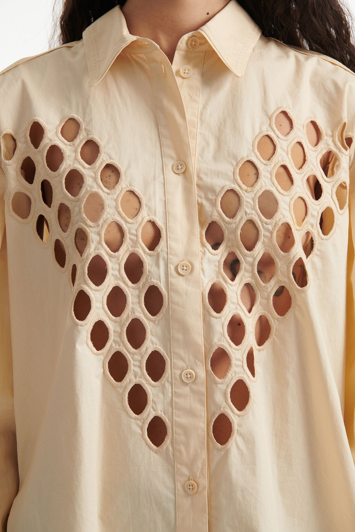 Female model wears off-white cotton shirt with see-through cutouts at chest and back. Model also wears brown wool pants and black high heels on white background. 