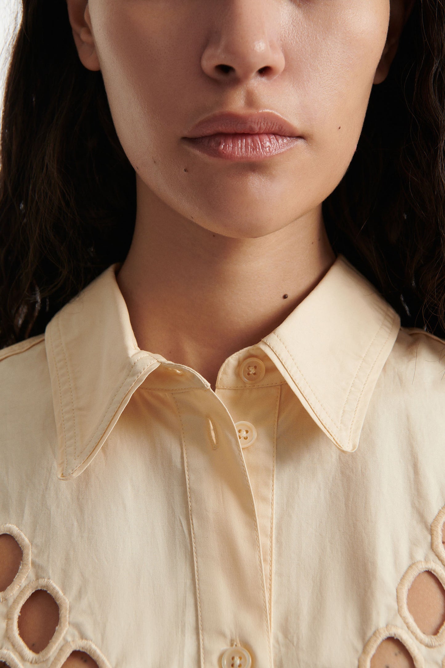 Female model wears off-white cotton shirt with see-through cutouts at chest and back. Model also wears brown wool pants and black high heels on white background. 