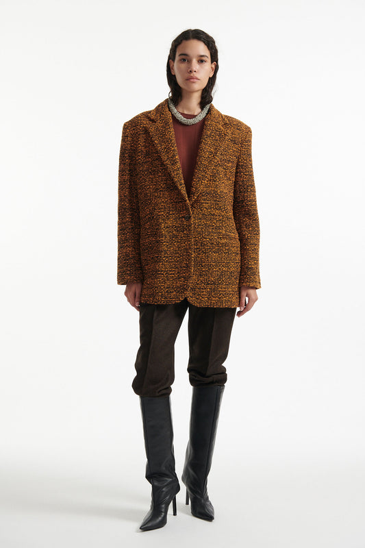 Female model wears orange and black tweed blazer, burnt orange cotton shirt, chunky gold necklace, brown straight leg trousers and black high heels on white background.