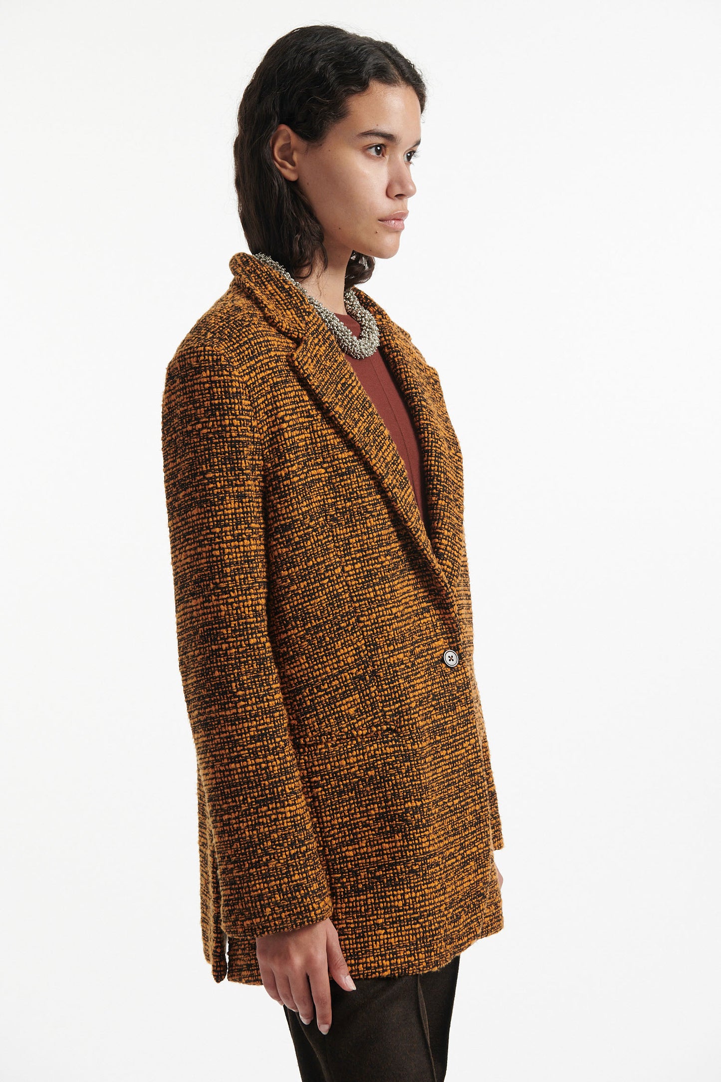 Female model wears orange and black tweed blazer, burnt orange cotton shirt, chunky gold necklace, brown straight leg trousers and black high heels on white background.