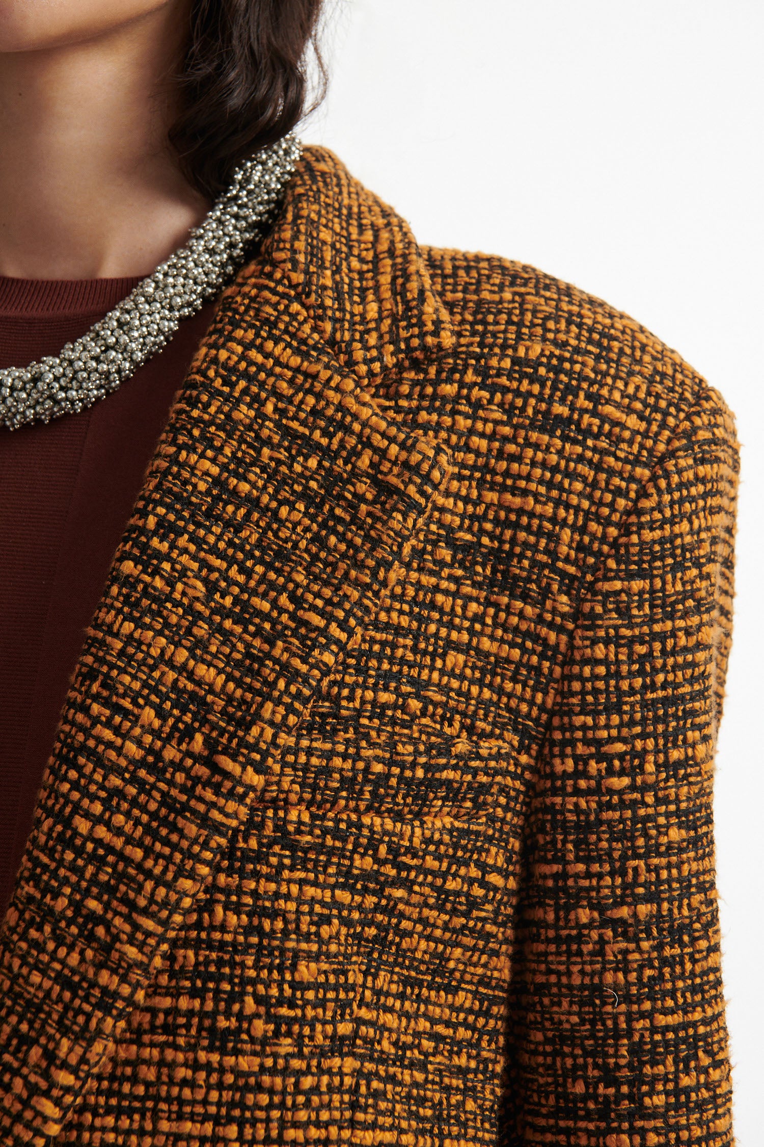 Female model wears orange and black tweed blazer, burnt orange cotton shirt, chunky gold necklace, brown straight leg trousers and black high heels on white background.