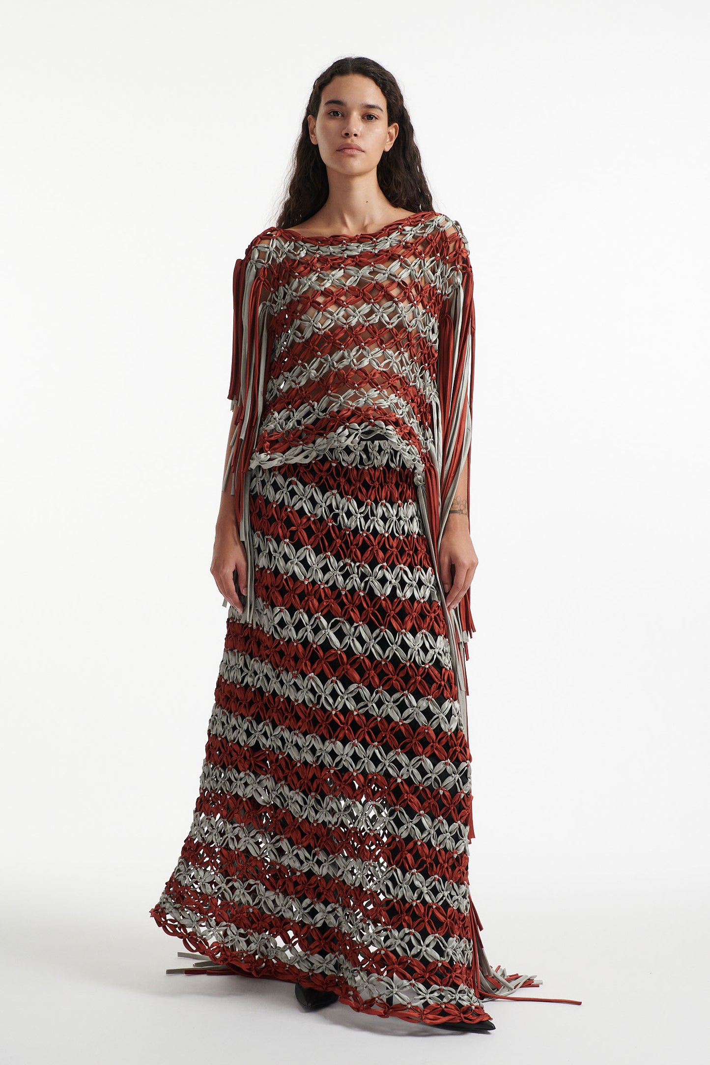 Female model wears knotted macrame set in burnt orange and sky grey. Both the top and skirt have asymmetric fringe hanging down either side. Model is on a white background. 