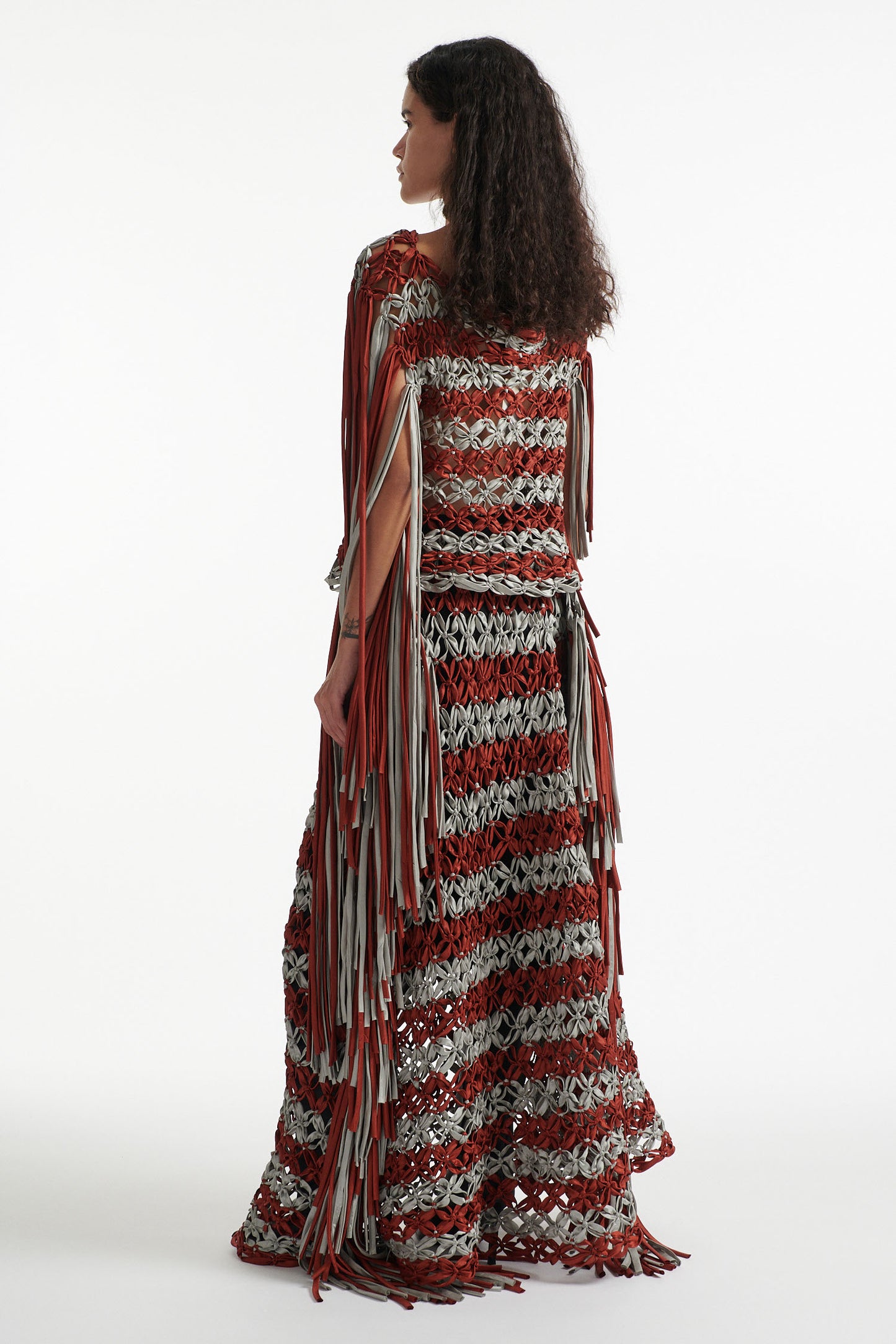 Female model wears knotted macrame set in burnt orange and sky grey. Both the top and skirt have asymmetric fringe hanging down either side. Model is on a white background. 