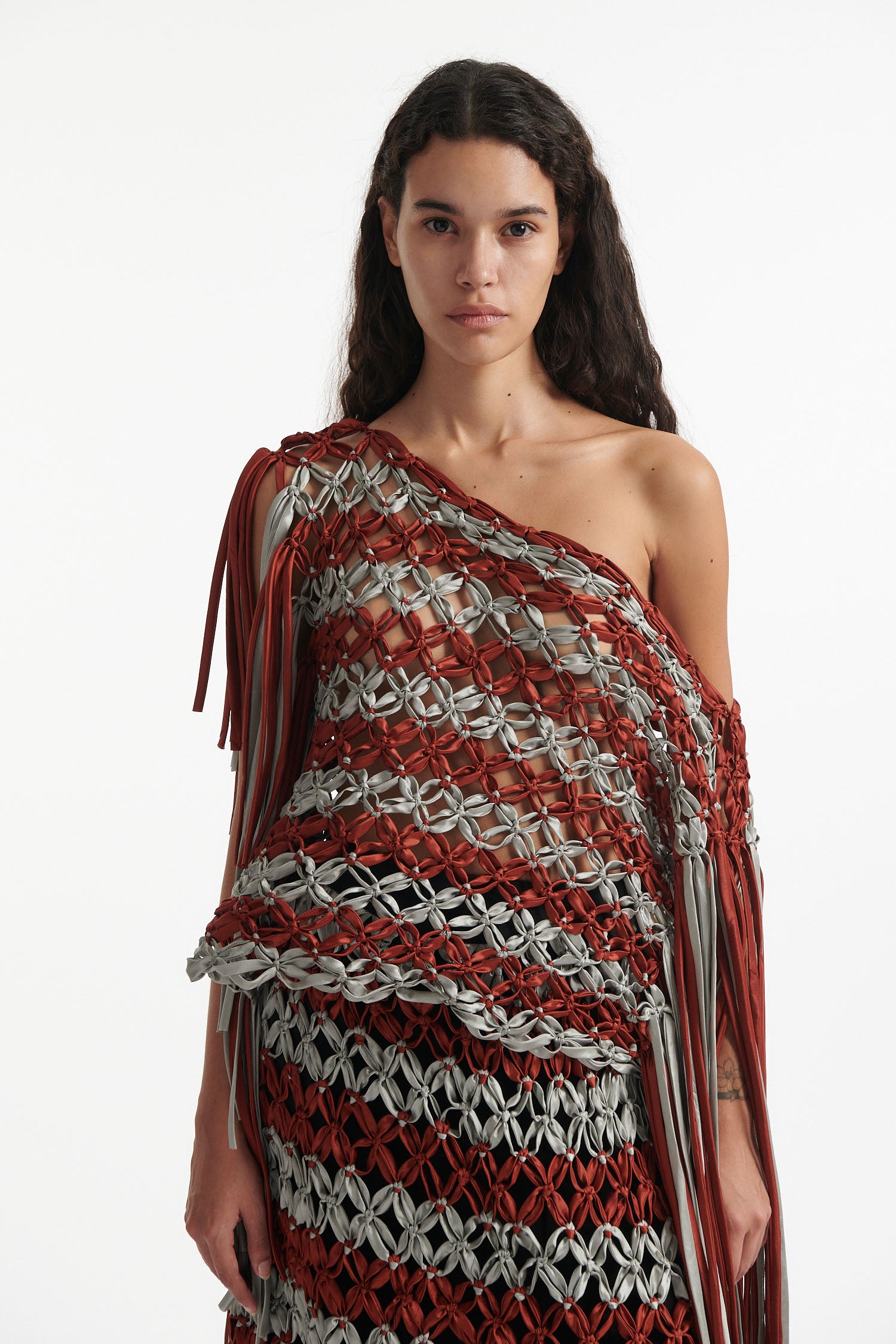 Female model wears knotted macrame set in burnt orange and sky grey. Both the top and skirt have asymmetric fringe hanging down either side. Model is on a white background. 