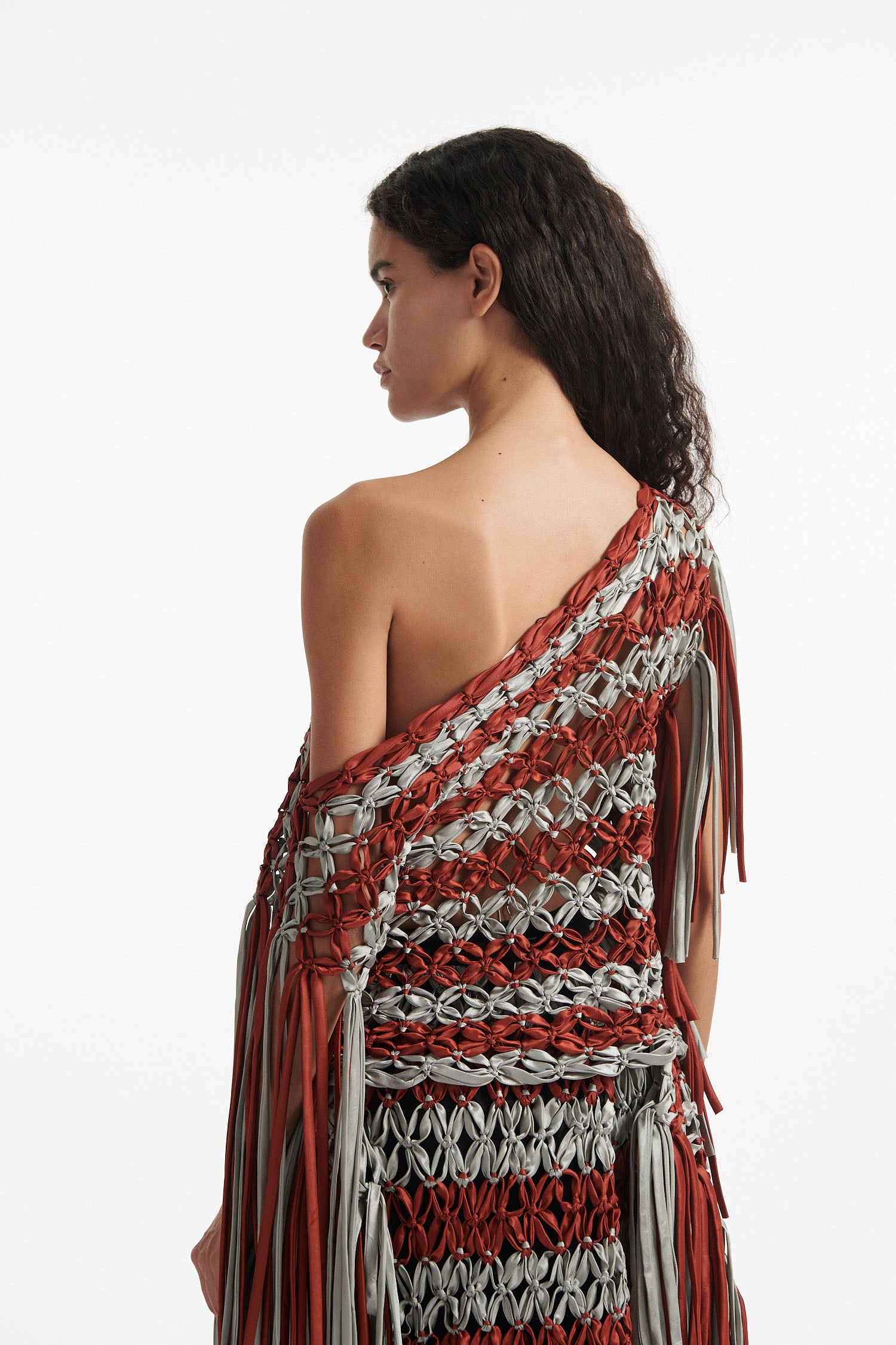 Female model wears knotted macrame set in burnt orange and sky grey. Both the top and skirt have asymmetric fringe hanging down either side. Model is on a white background. 