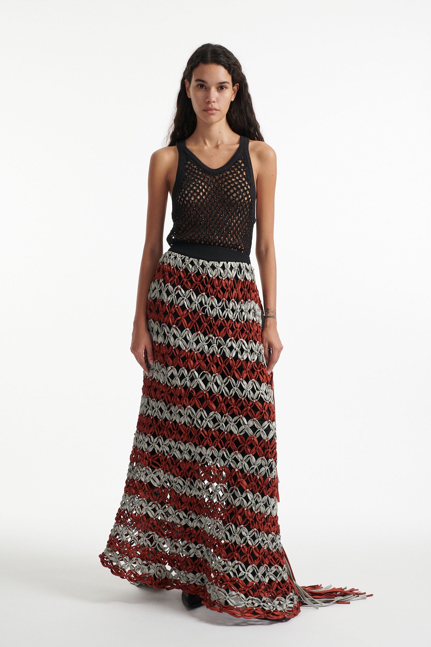 Female model wears black crystal embellished top with knotted macrame skirt in burnt orange and sky grey with fringe hanging down either side. Model is on a white background. 