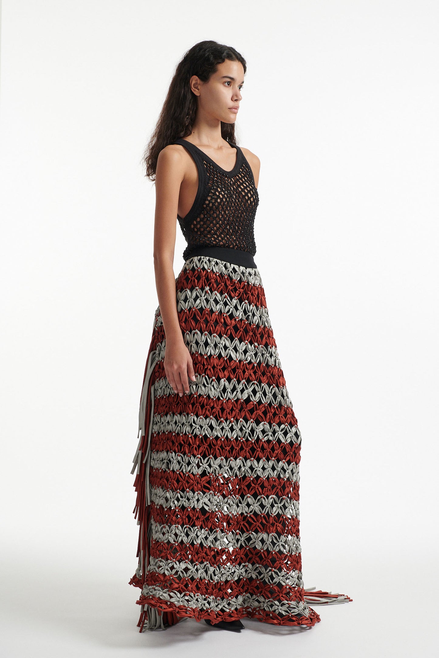 Female model wears black crystal embellished top with knotted macrame skirt in burnt orange and sky grey with fringe hanging down either side. Model is on a white background. 