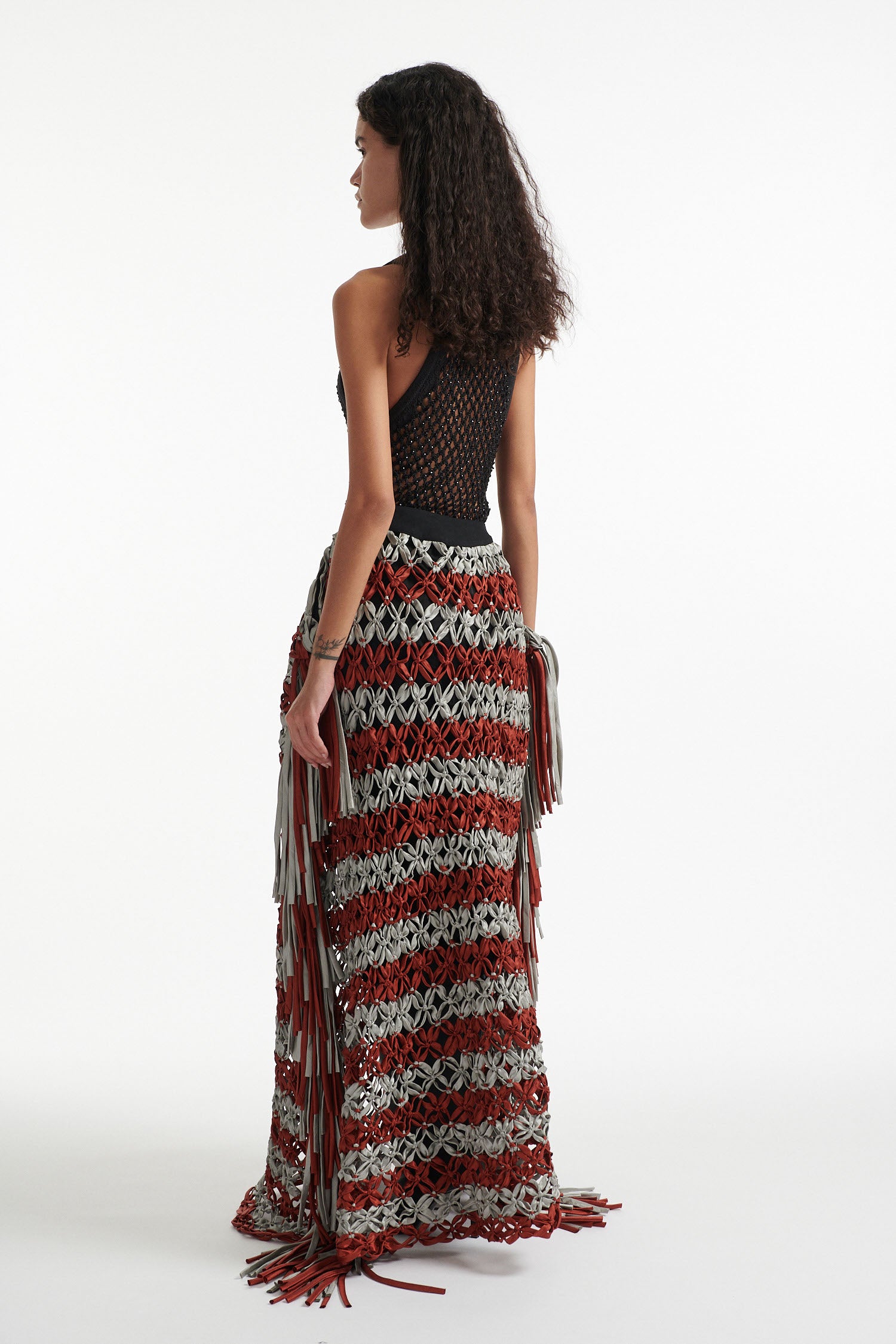 Female model wears black crystal embellished top with knotted macrame skirt in burnt orange and sky grey with fringe hanging down either side. Model is on a white background. 