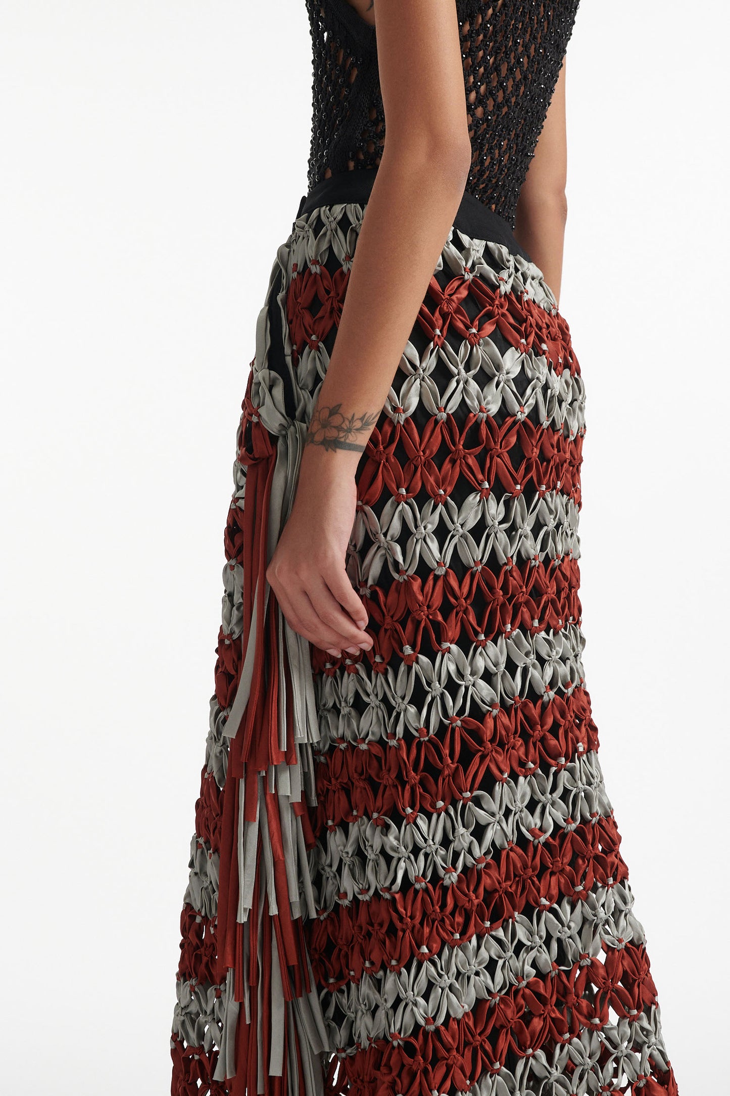 Female model wears black crystal embellished top with knotted macrame skirt in burnt orange and sky grey with fringe hanging down either side. Model is on a white background. 