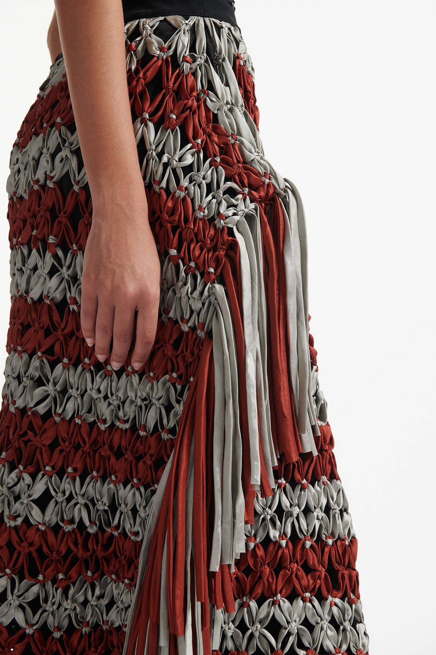 Female model wears black crystal embellished top with knotted macrame skirt in burnt orange and sky grey with fringe hanging down either side. Model is on a white background. 