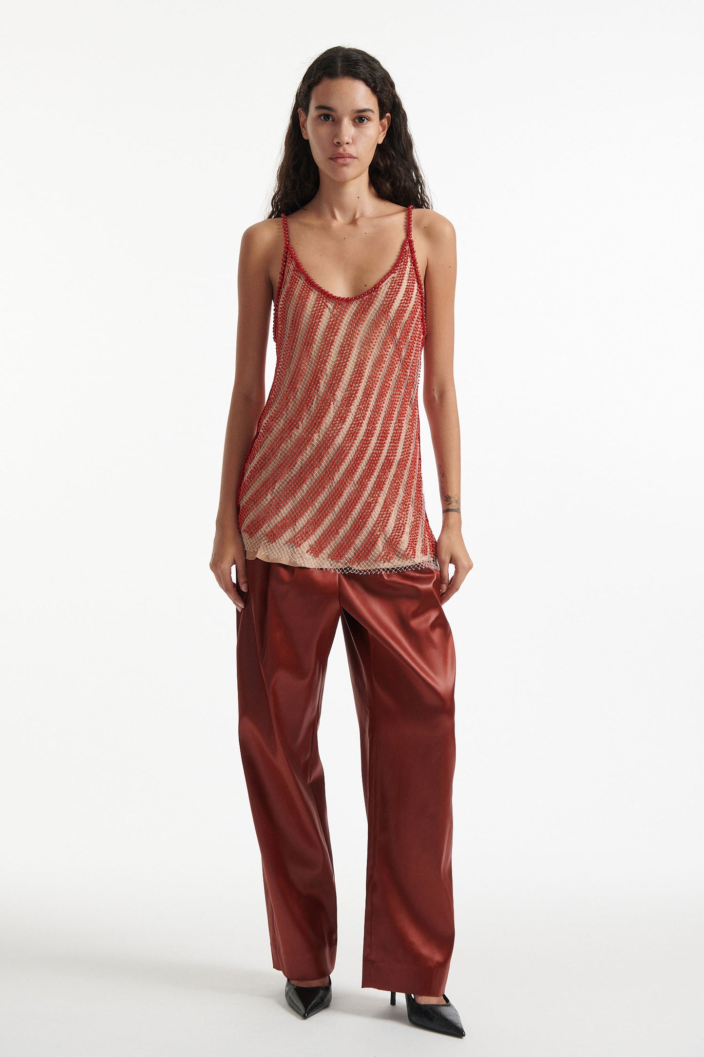 Female model wears diagonal beaded red and tan camisole  over burnt orange trousers with black high heels on white background. 