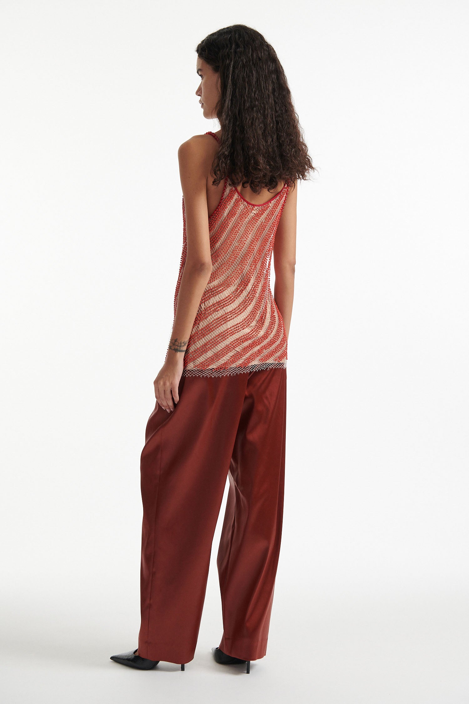 Female model wears diagonal beaded red and tan camisole  over burnt orange trousers with black high heels on white background. 