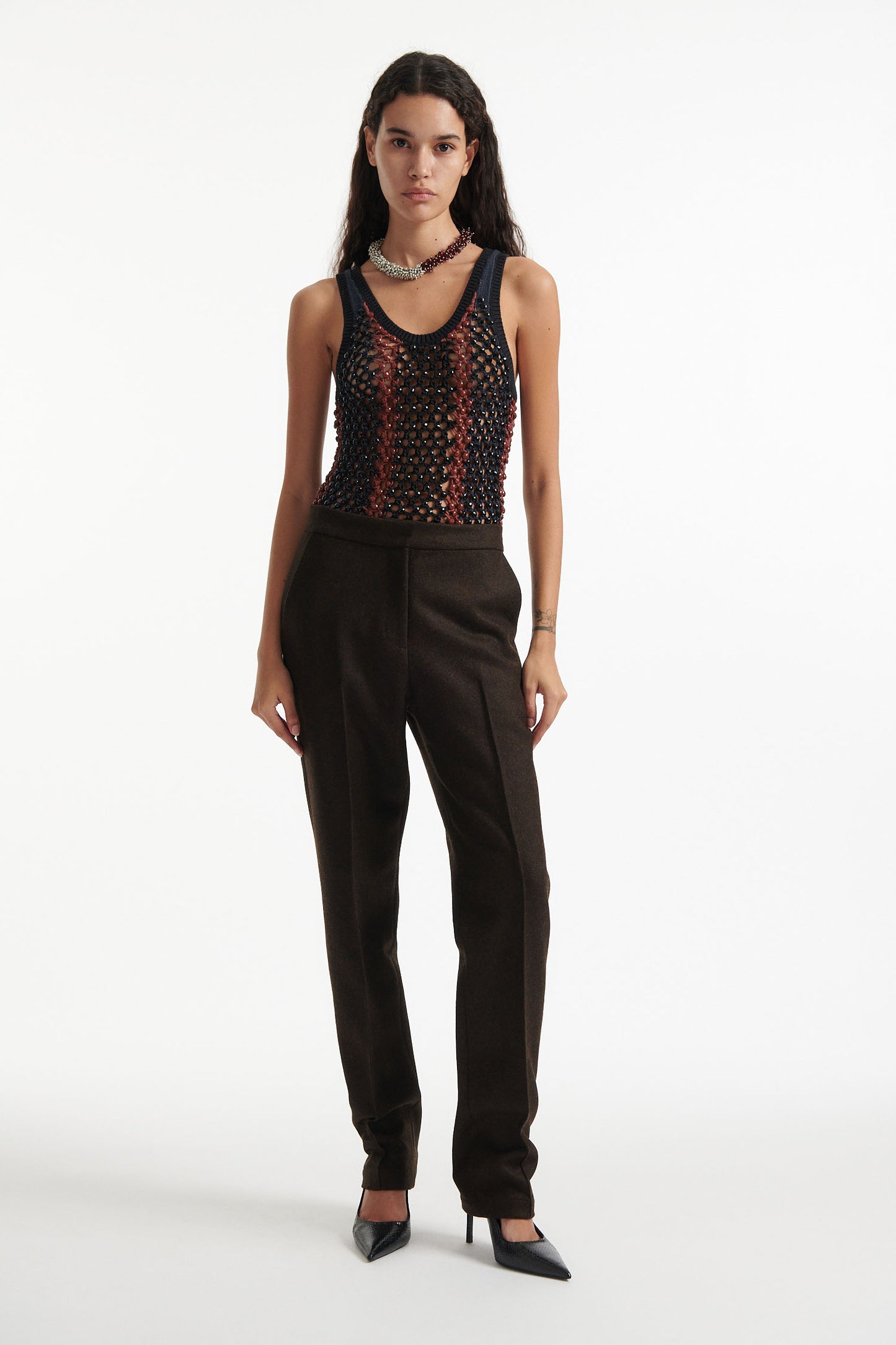 Female model wears striped crystal embellished tank top with slim brown wool trousers and black high heels on white background.