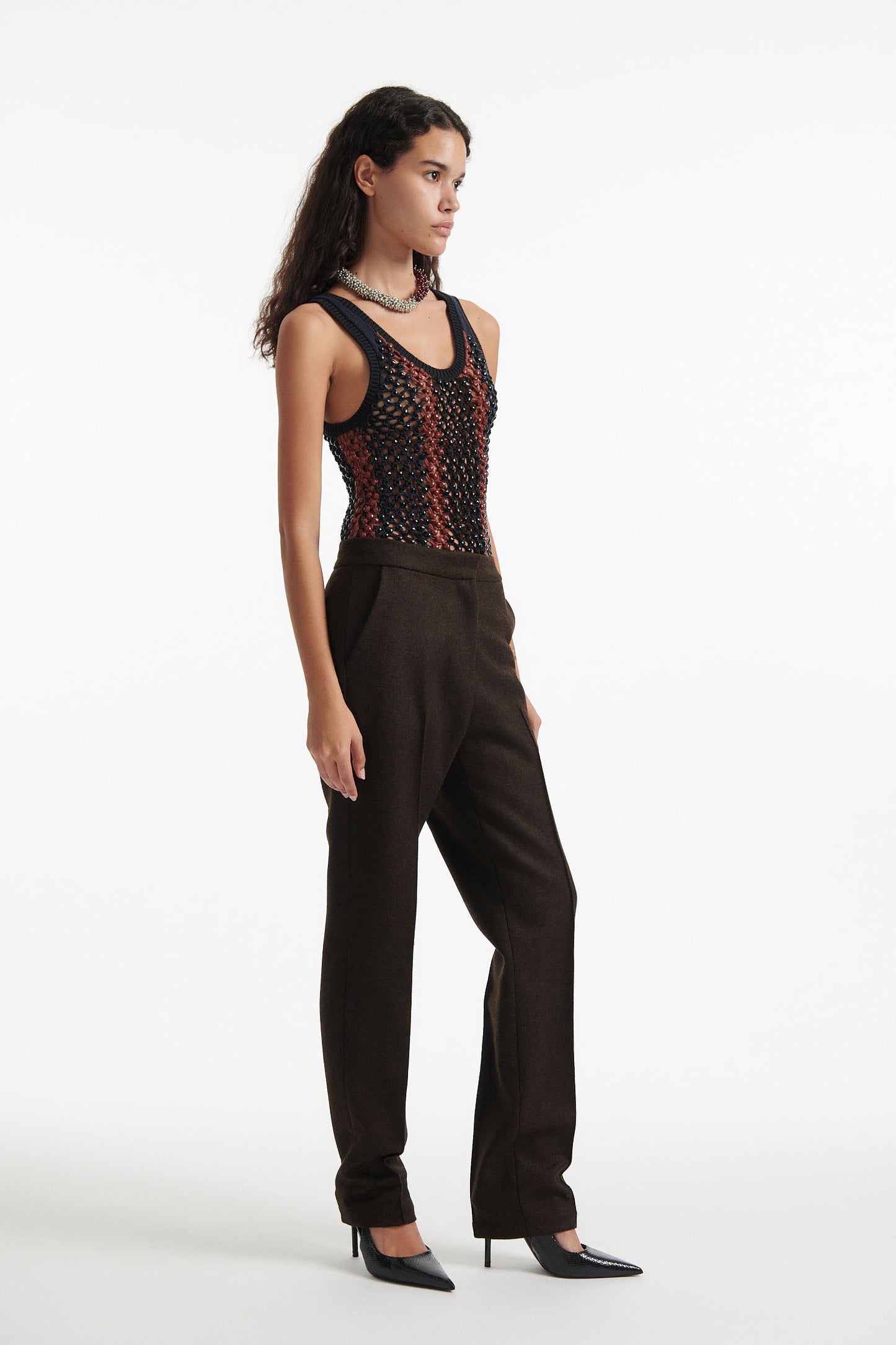 Female model wears striped crystal embellished tank top with slim brown wool trousers and black high heels on white background.