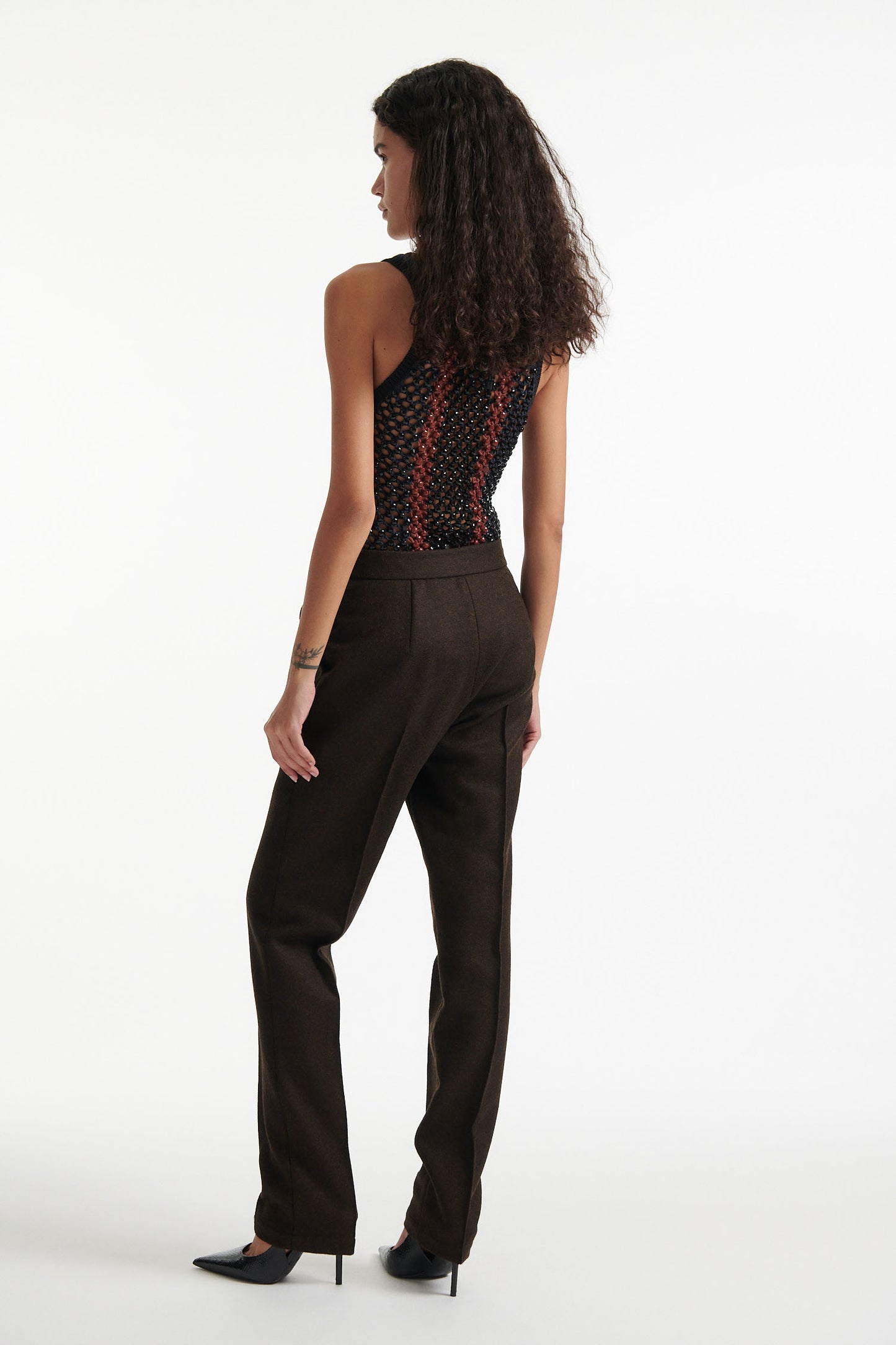 Female model wears striped crystal embellished tank top with slim brown wool trousers and black high heels on white background.