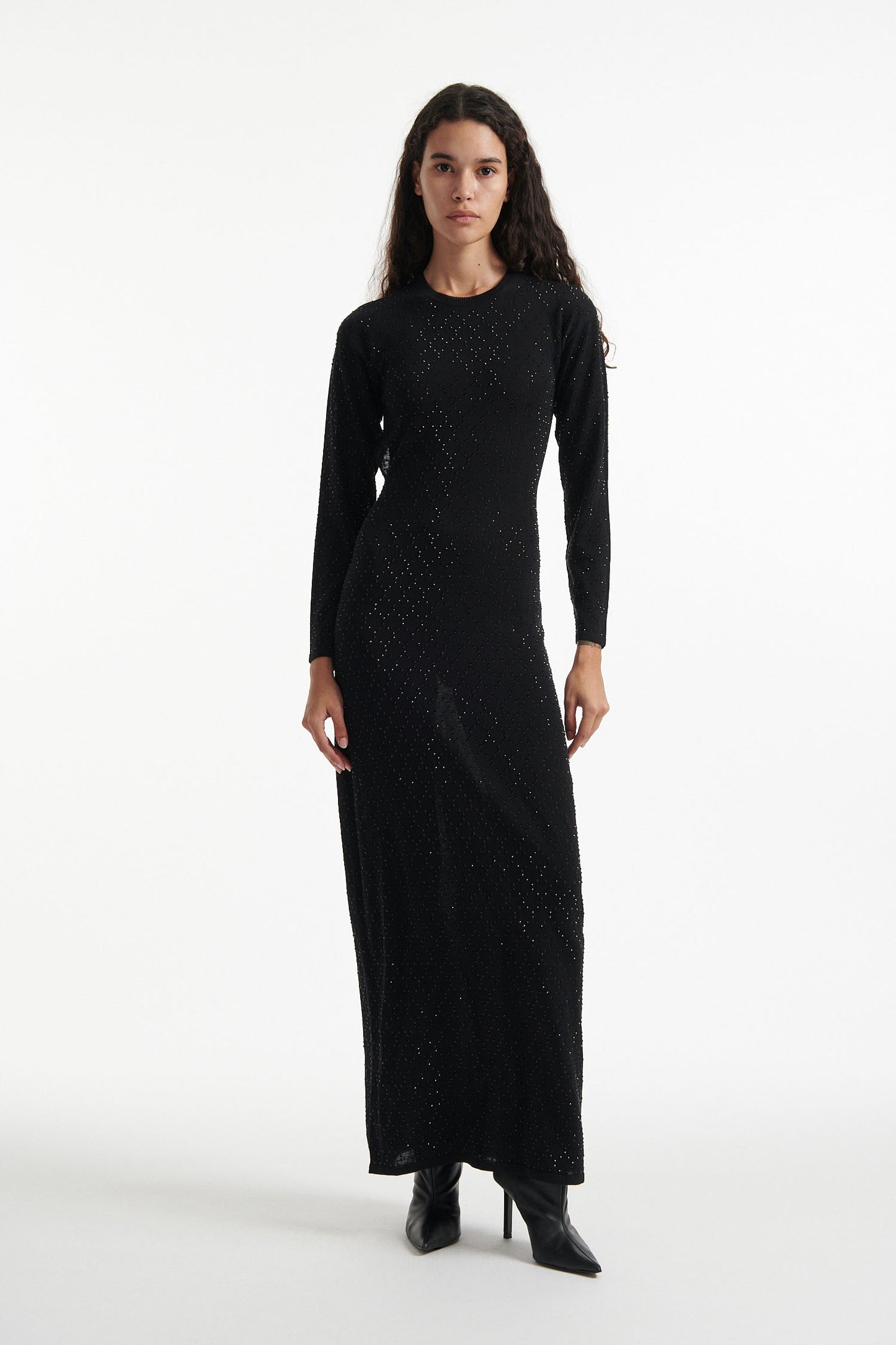 Female model wears open-back crystal embellished long sleeve black dress and black high heels on white background. 