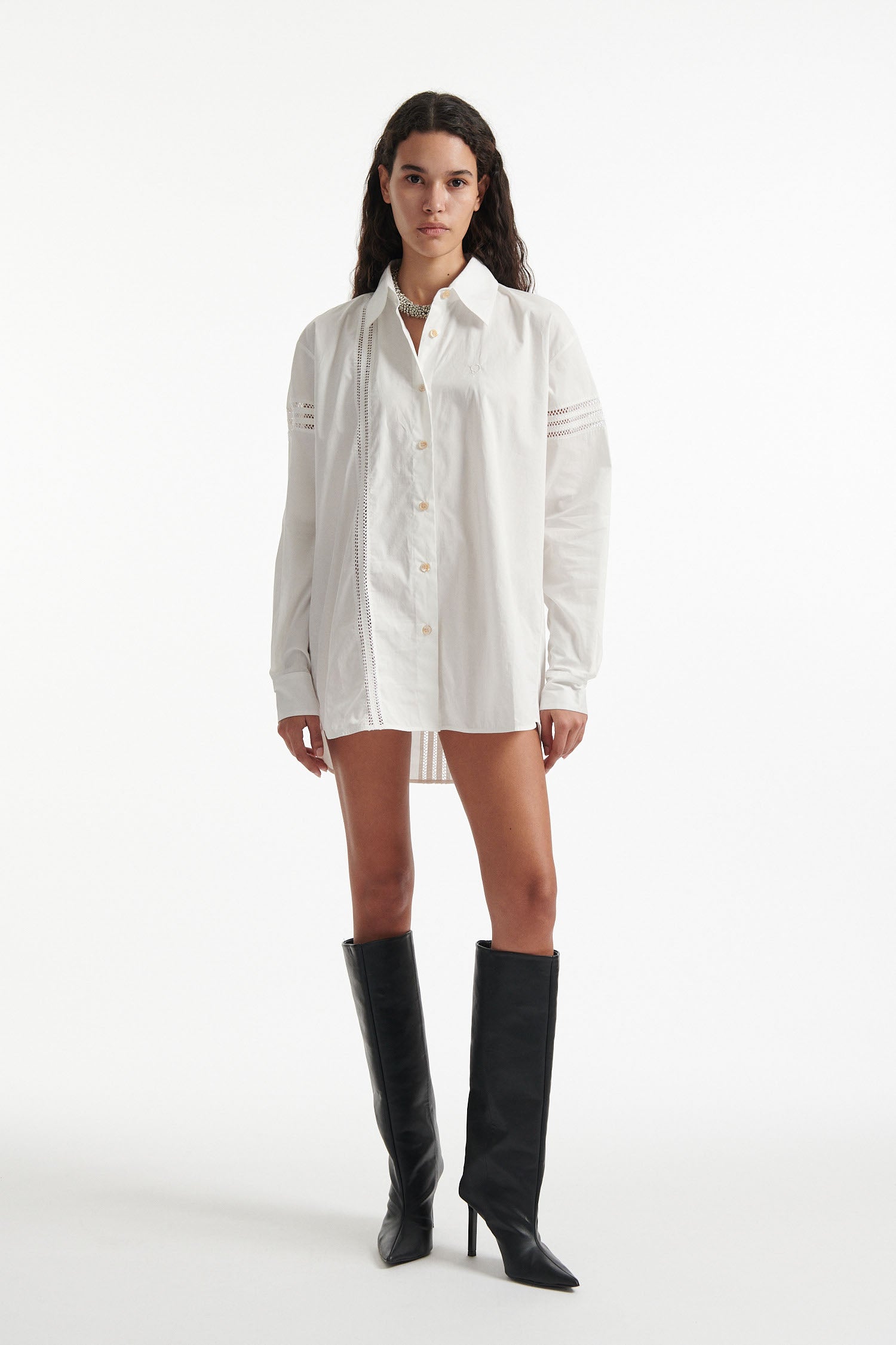 Female model wears white poplin shirt with exposed stitches and black high heel boots on white background. 