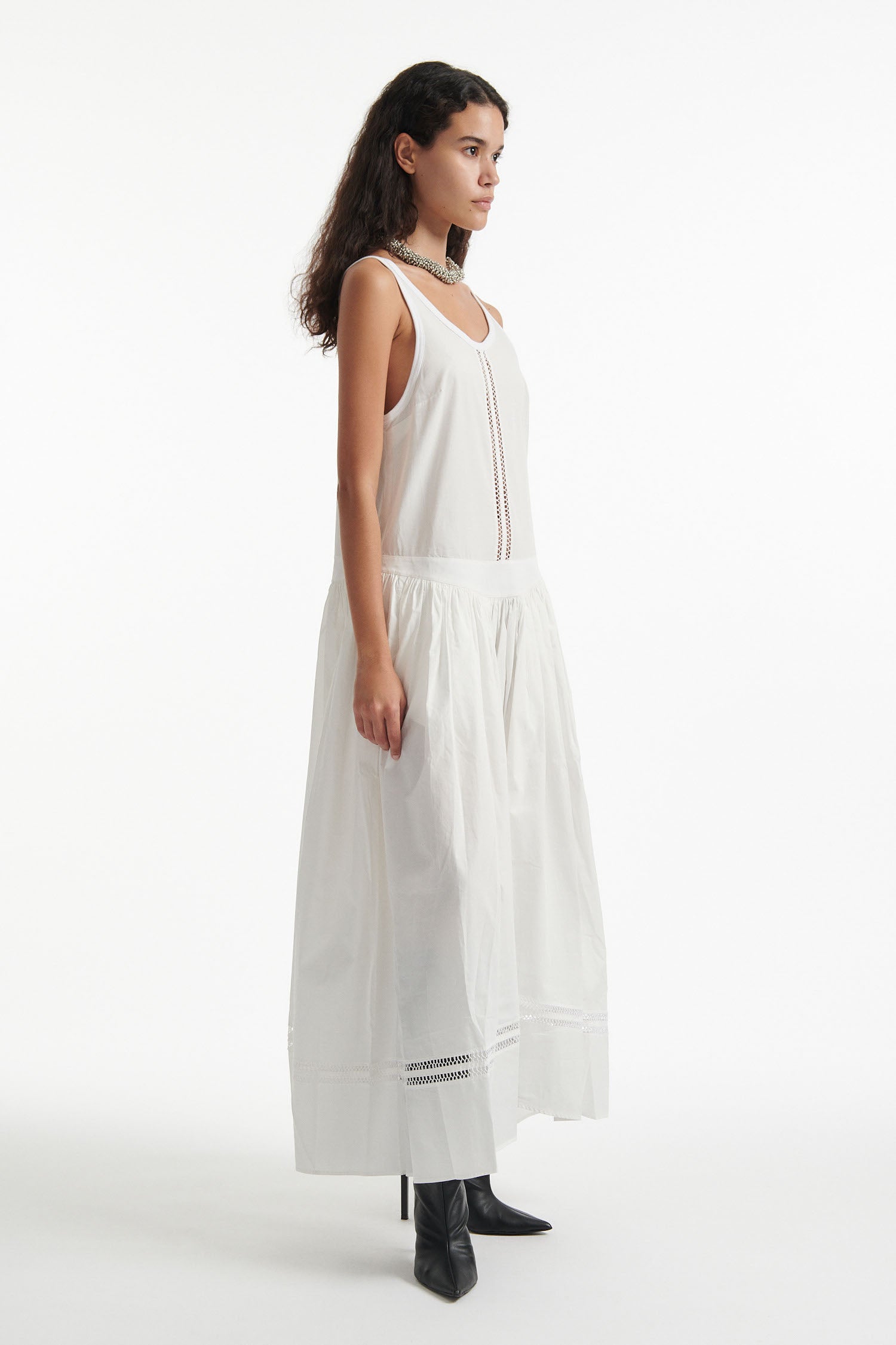 Female model wears white poplin sleeveless dress with cutout stitched detailing and black high heels on white background. 