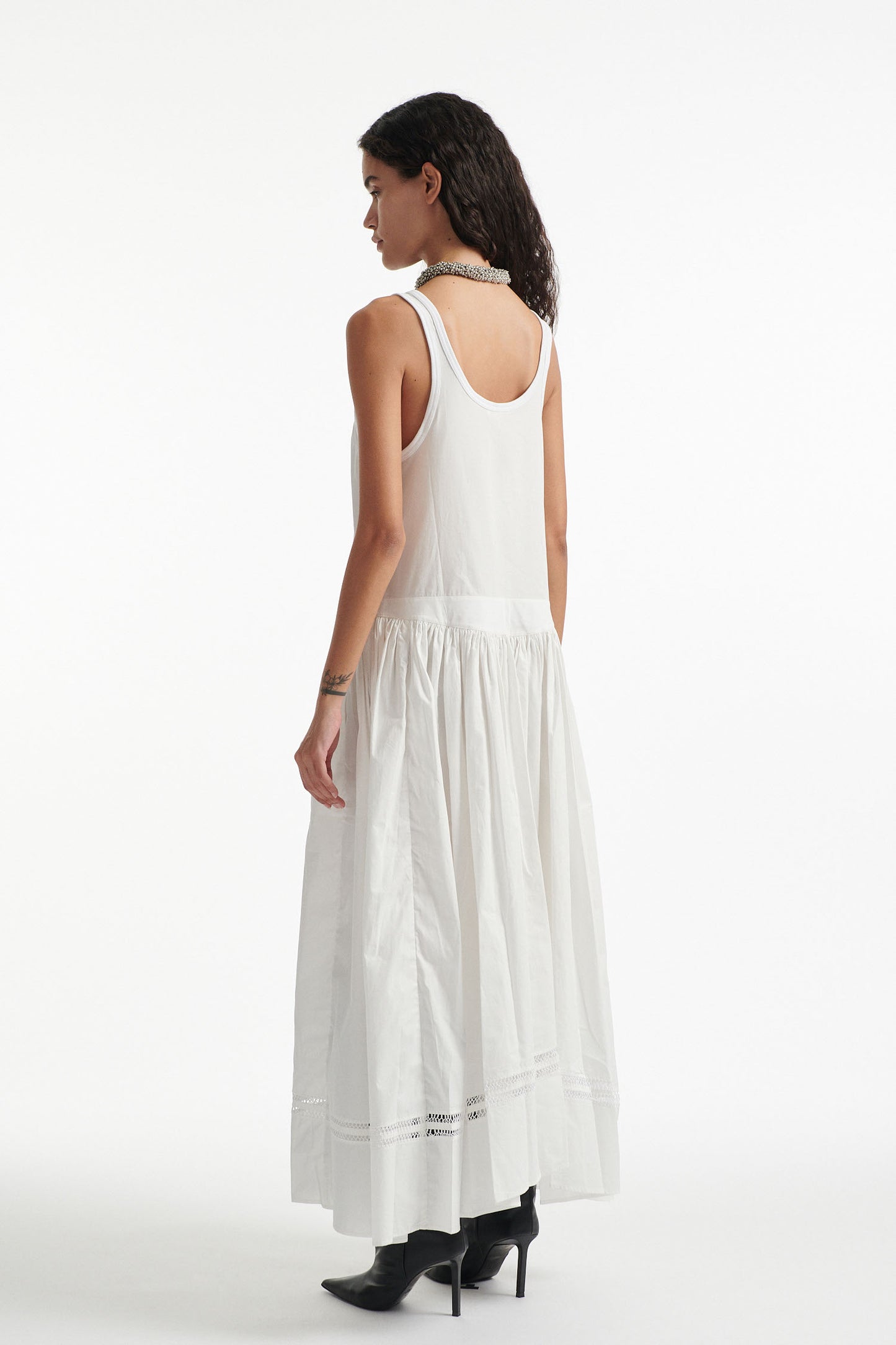 Female model wears white poplin sleeveless dress with cutout stitched detailing and black high heels on white background. 