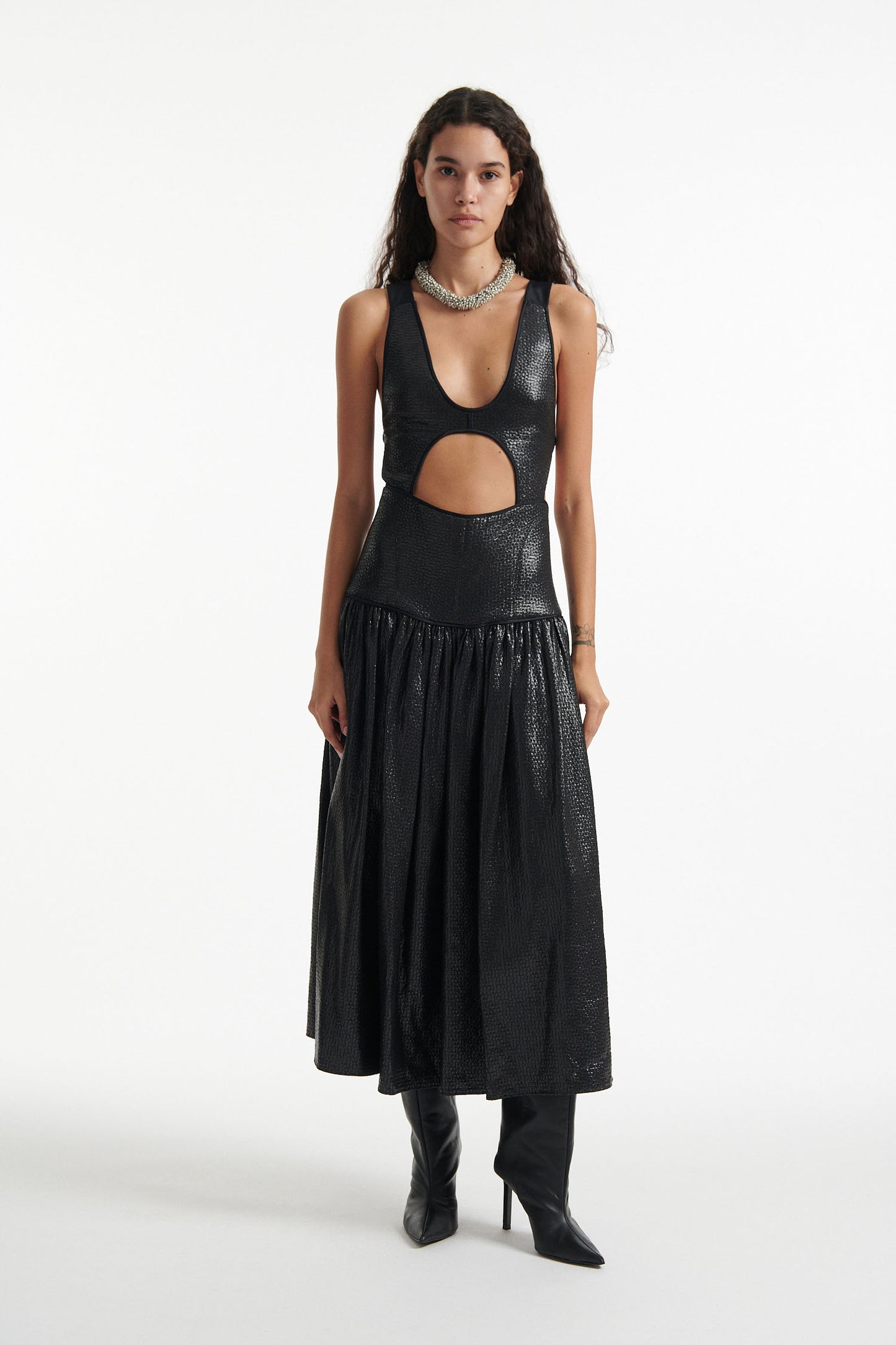 Female model wears open-back black sleeveless dress and black high heels on white background. Dress has cutout in the center.