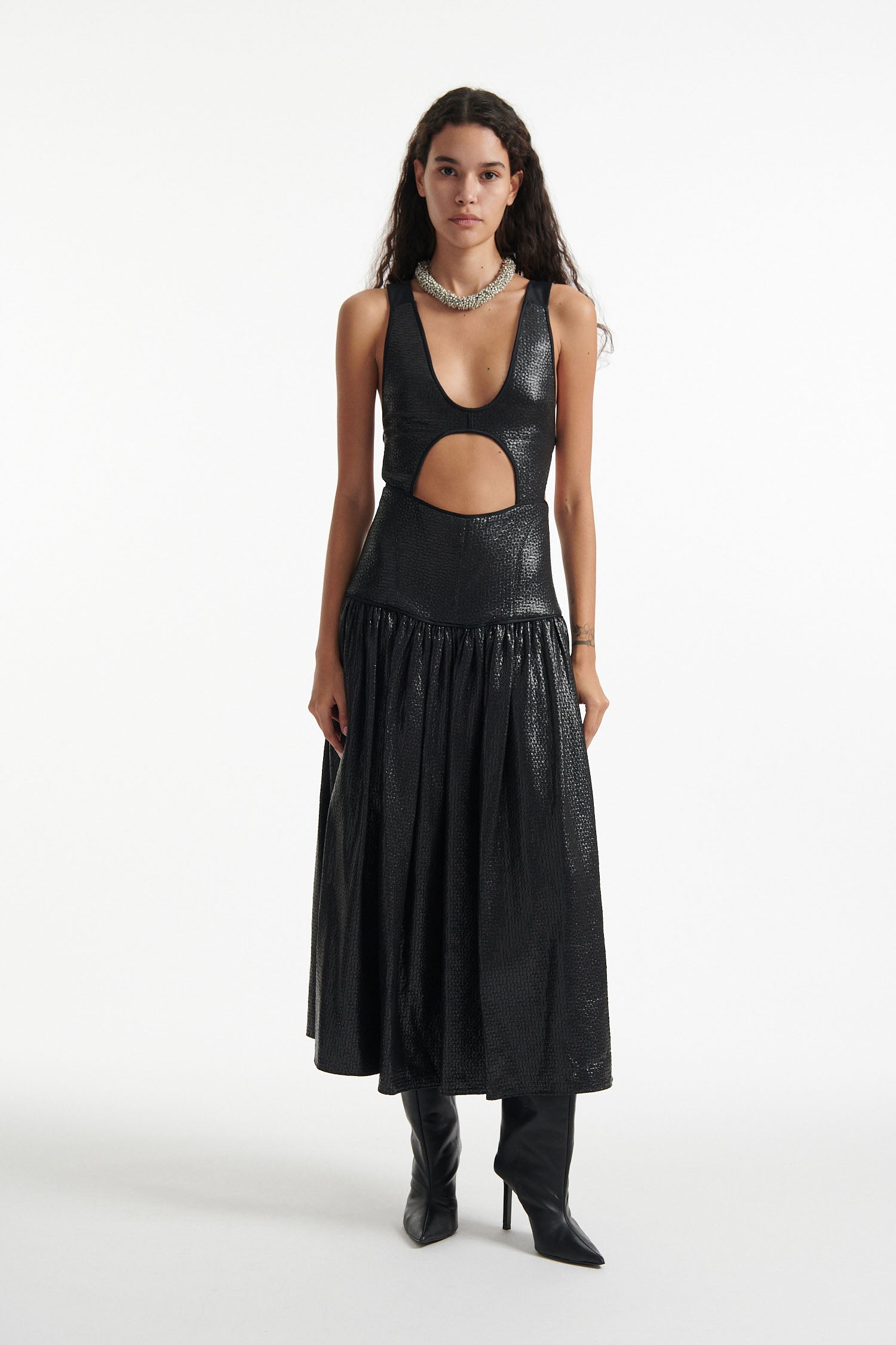 Female model wears open-back black sleeveless dress and black high heels on white background. Dress has cutout in the center.