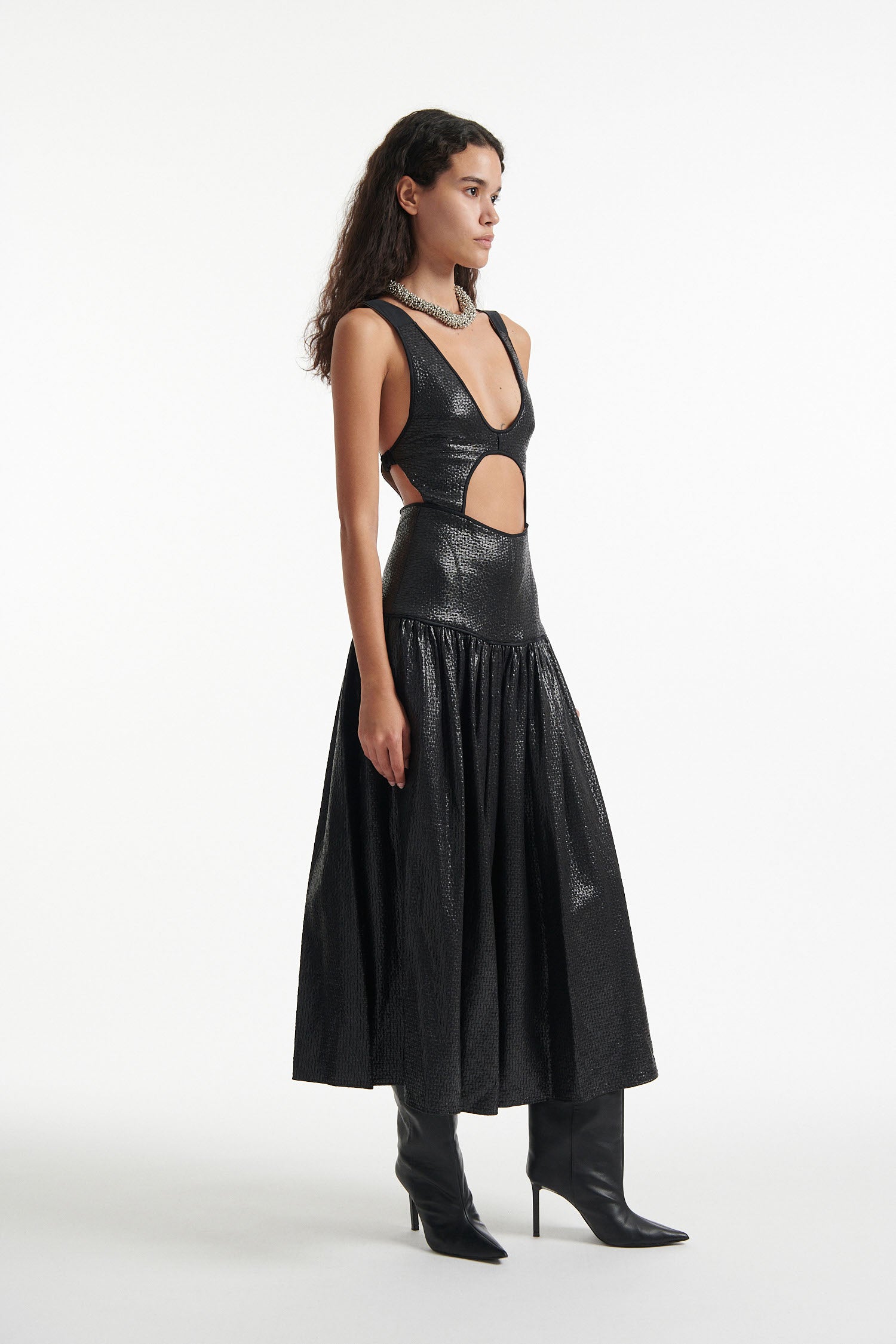 Female model wears open-back black sleeveless dress and black high heels on white background. Dress has cutout in the center.
