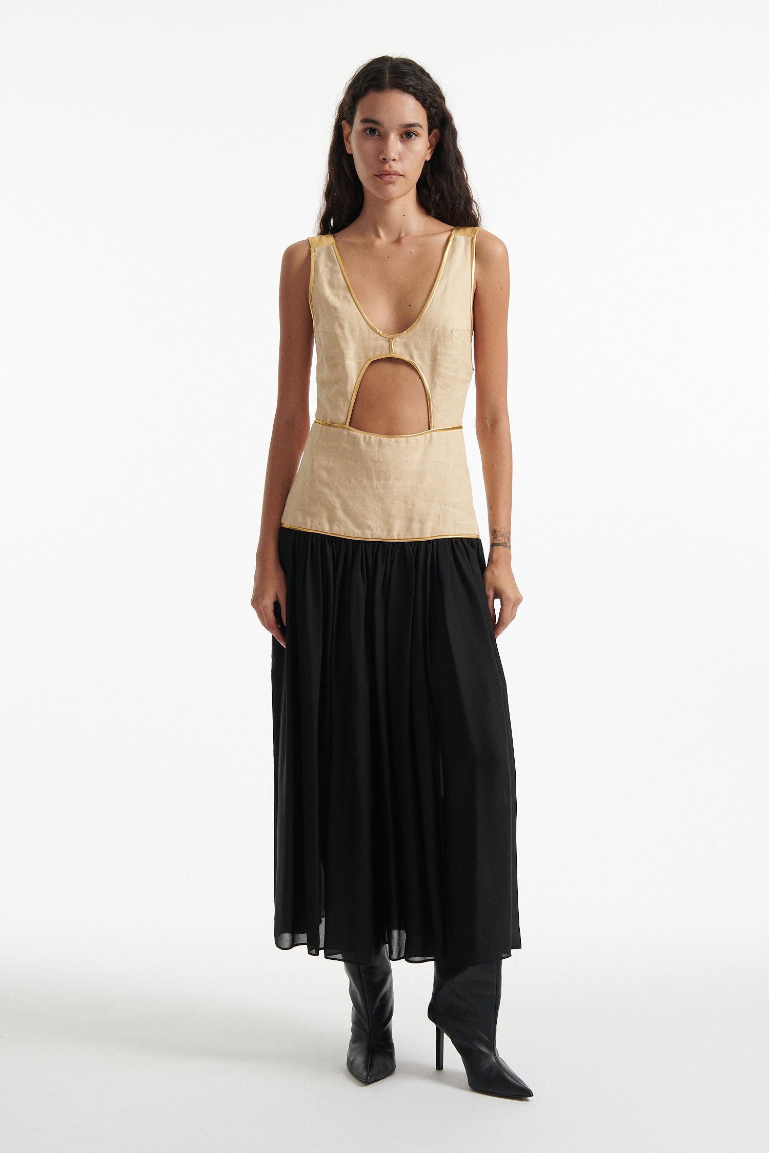 Female model wears sleeveless dress with beige upper and black lower half. The whole dress has gold piping and the model is wearing black high heels on white background. 