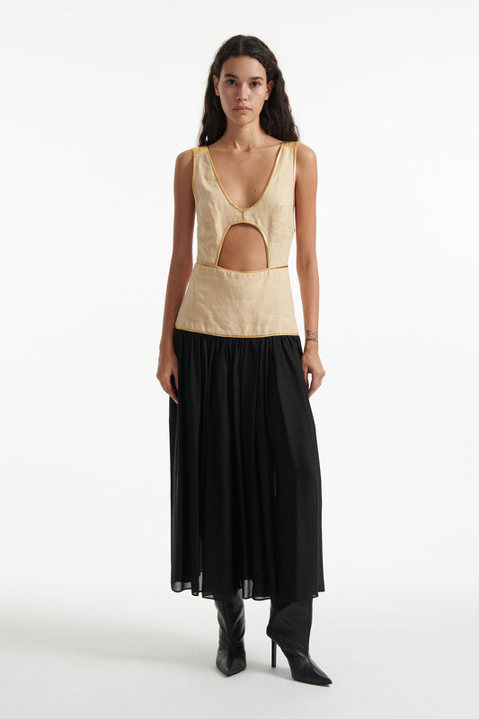 Female model wears sleeveless dress with beige upper and black lower half. The whole dress has gold piping and the model is wearing black high heels on white background. 