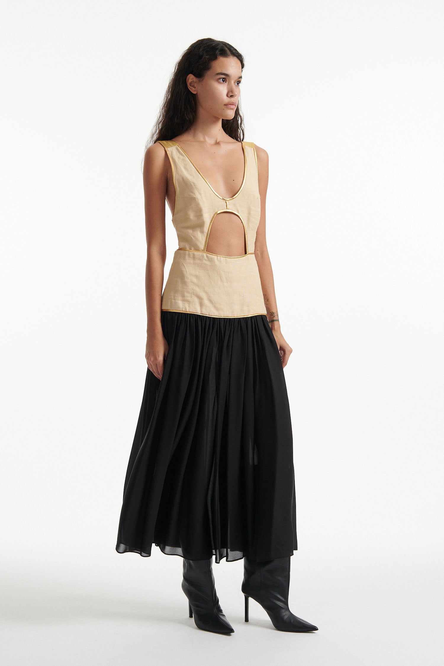 Female model wears sleeveless dress with beige upper and black lower half. The whole dress has gold piping and the model is wearing black high heels on white background. 