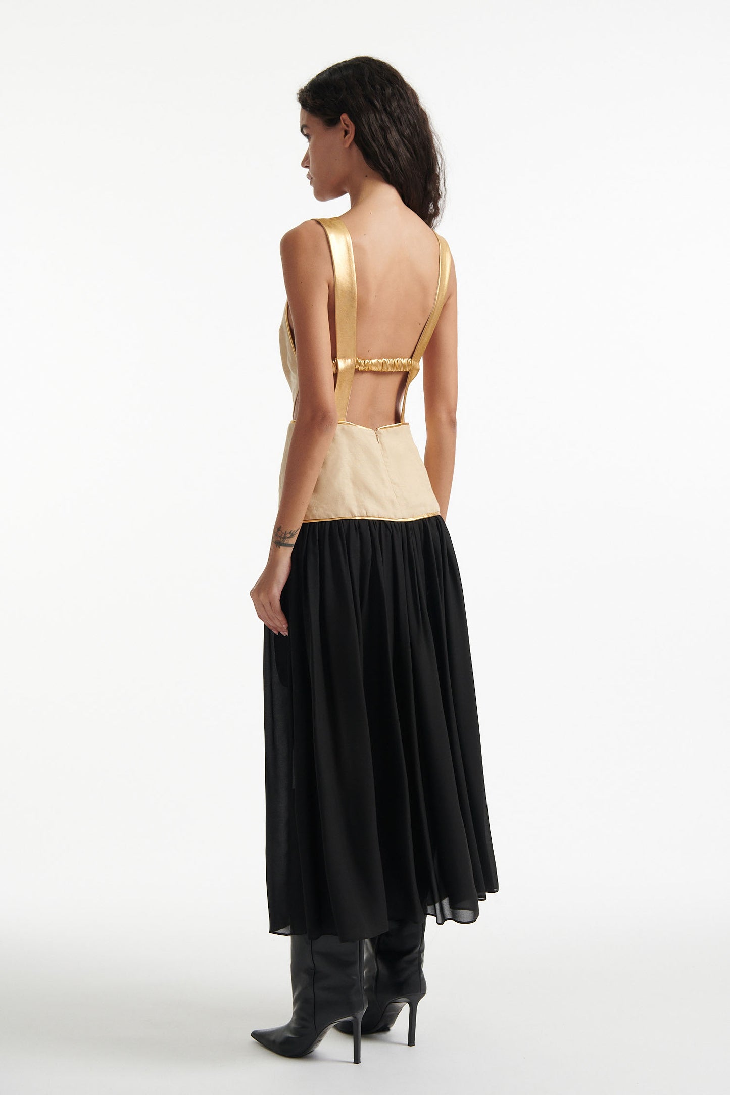Female model wears open-black sleeveless dress with beige upper and black lower half. The whole dress has gold piping and the model is wearing black high heels on white background. 
