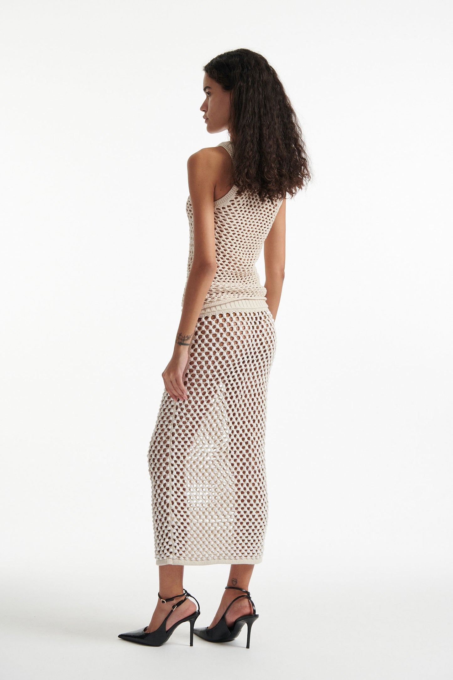 Female model wears beige mesh tank top and white crystal embellished skirt with black high heels on white background. 