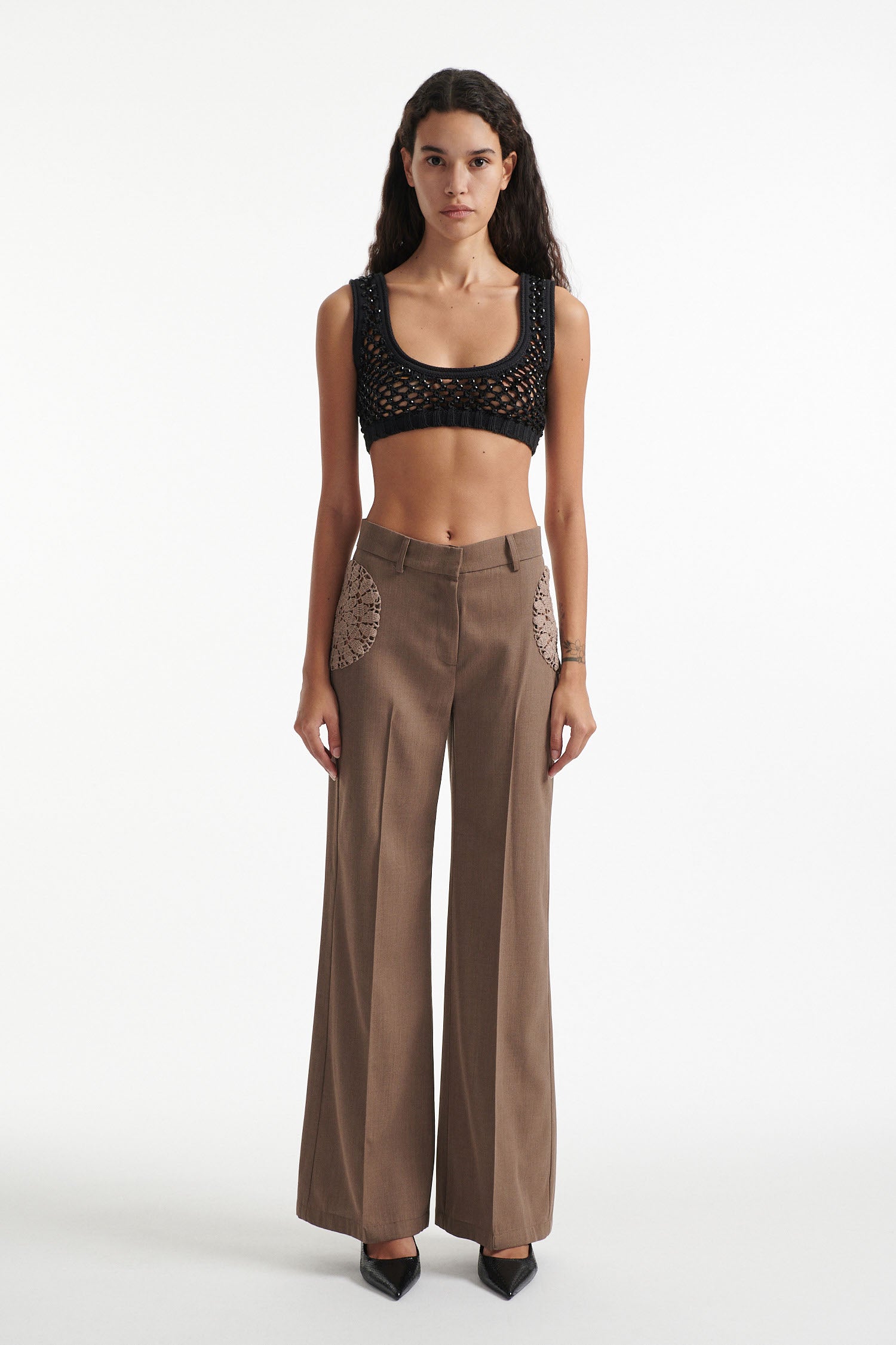 Female model wears brown trousers with crochet inserts and crystal embellished bralette with black high heels on white background. 