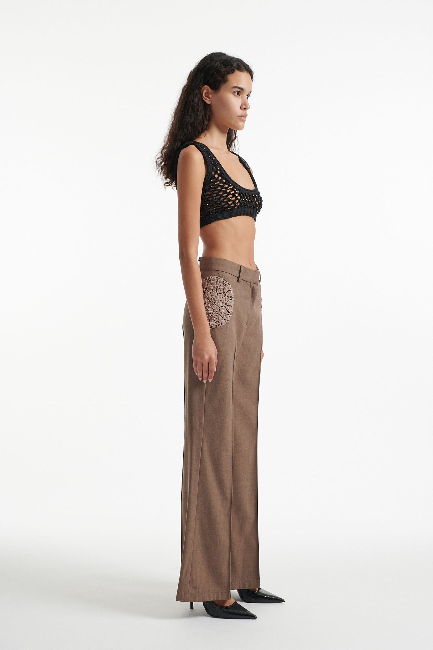 Female model wears brown trousers with crochet inserts and crystal embellished bralette with black high heels on white background. 