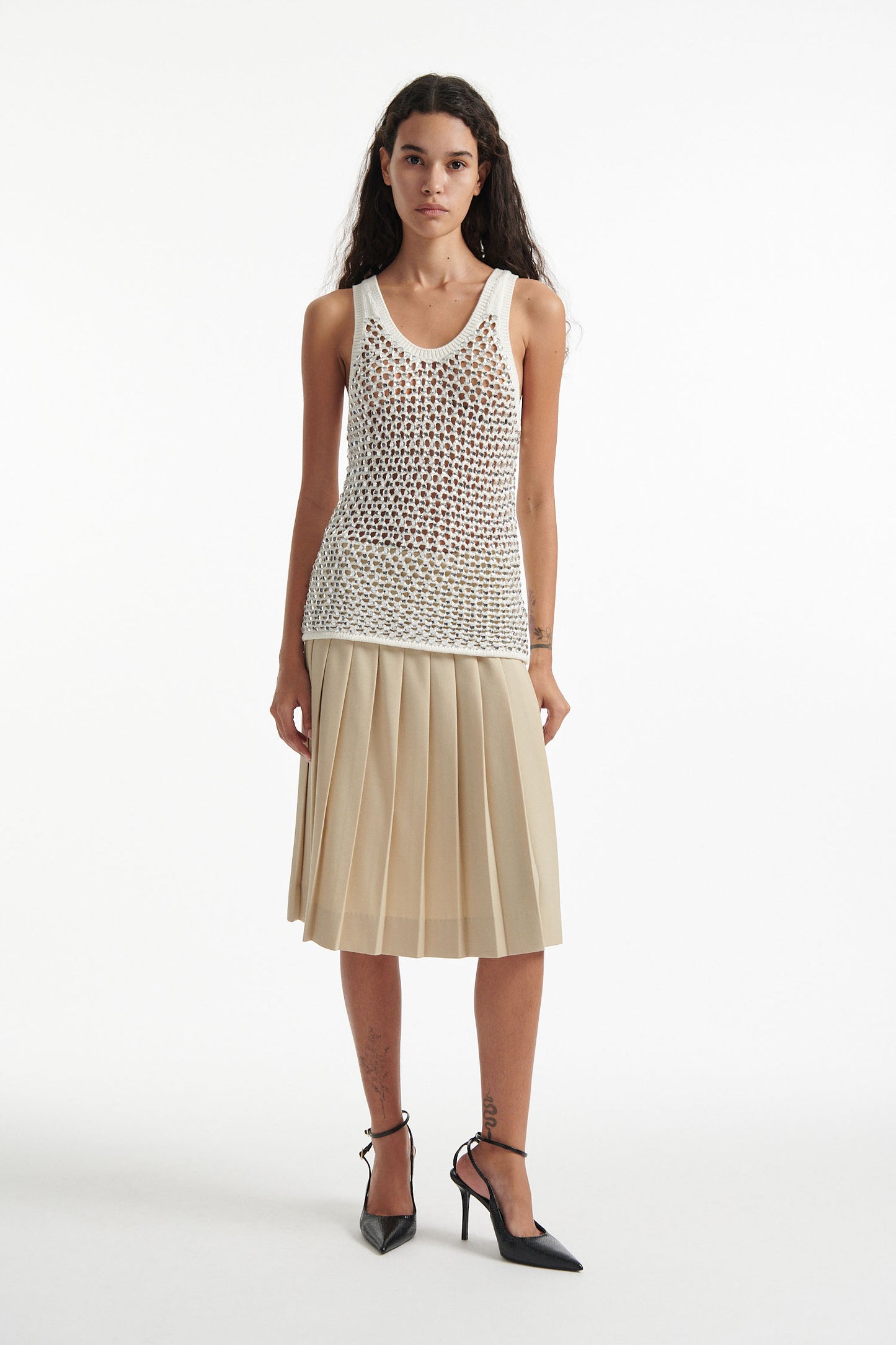 Female model wears white crystal embellished tank with beige pleated skirt and black high heels on white background.