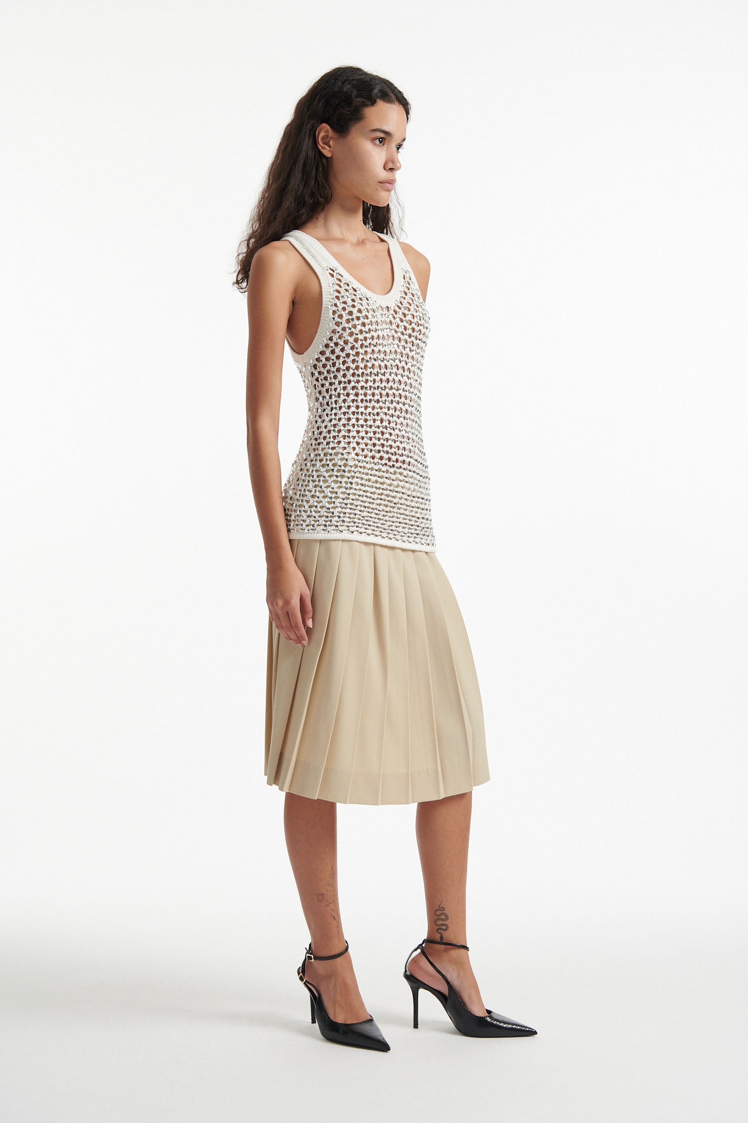 Female model wears white crystal embellished tank with beige pleated skirt and black high heels on white background.