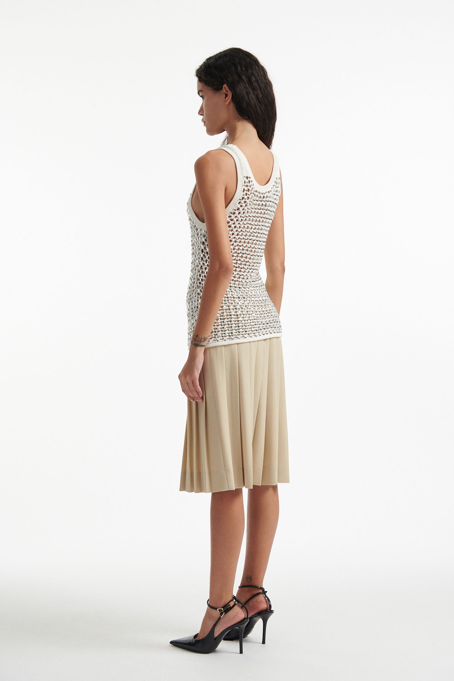 Female model wears white crystal embellished tank with beige pleated skirt and black high heels on white background.