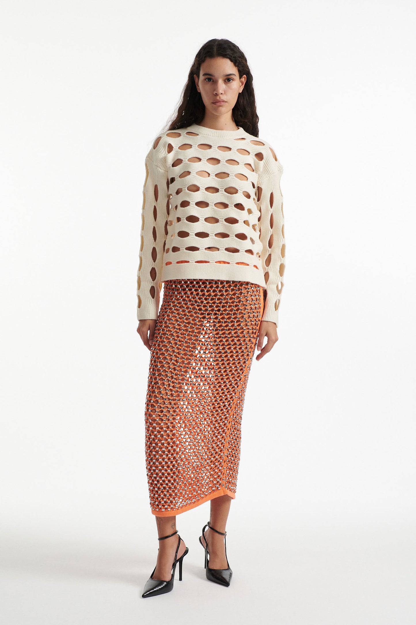 Female model wears open knit off-white long sleeve crewneck with crystal embellished bright orange skirt and black high heels on white background. 