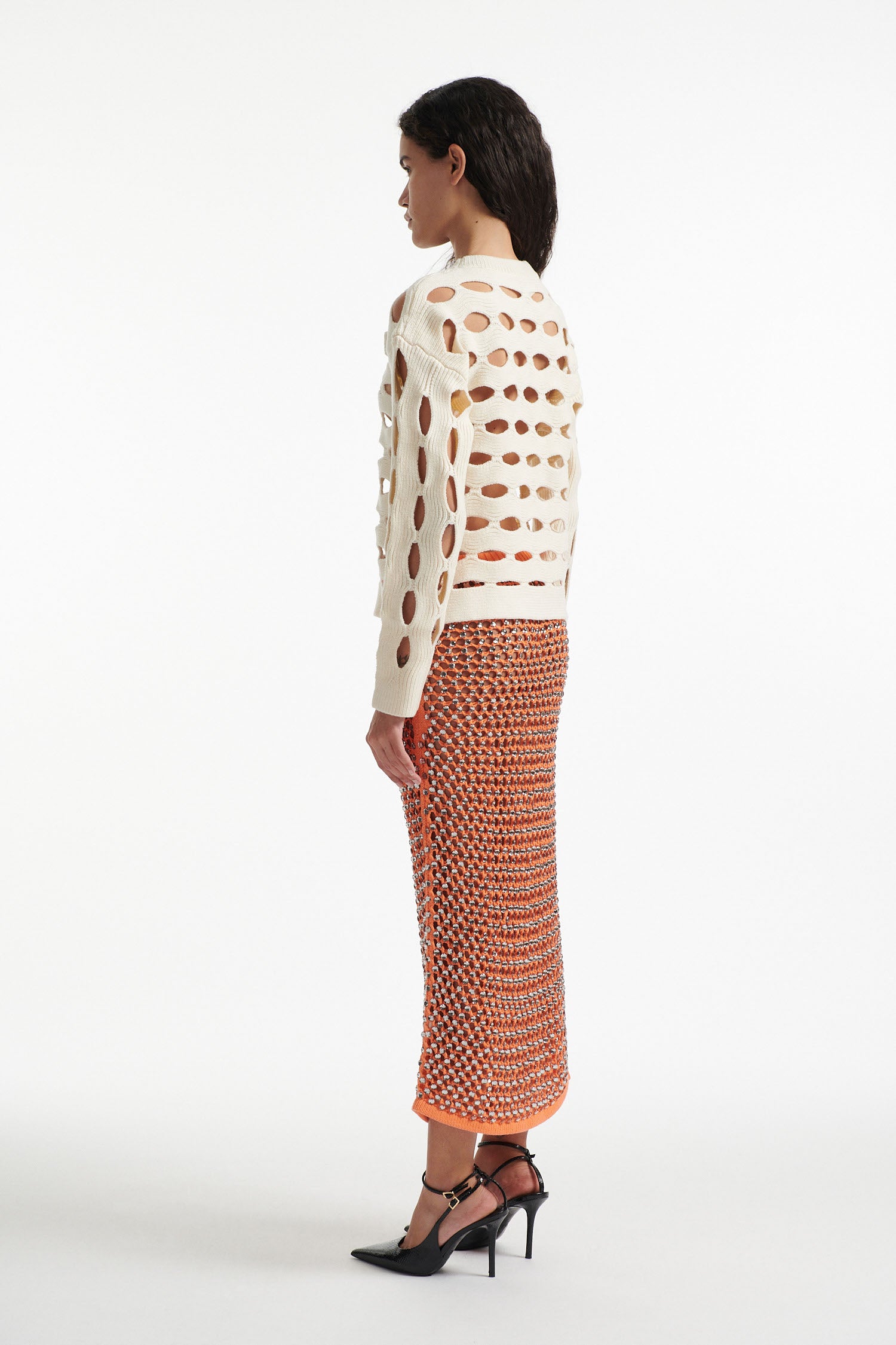 Female model wears open knit off-white long sleeve crewneck with crystal embellished bright orange skirt and black high heels on white background. 