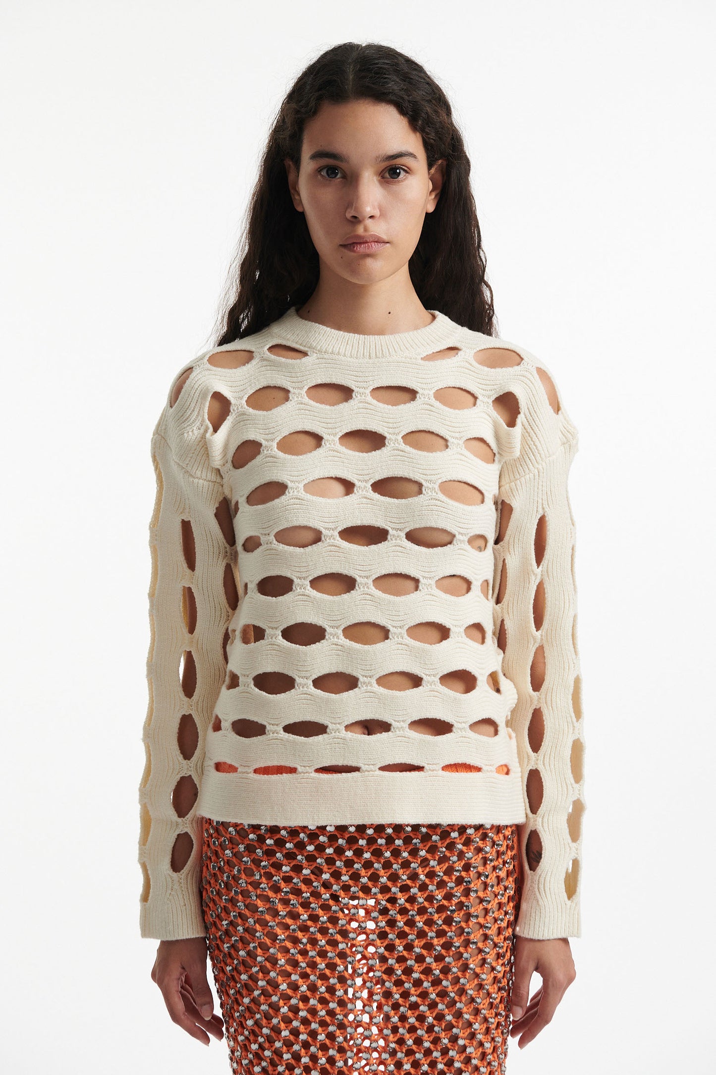 Female model wears open knit off-white long sleeve crewneck with crystal embellished bright orange skirt and black high heels on white background. 