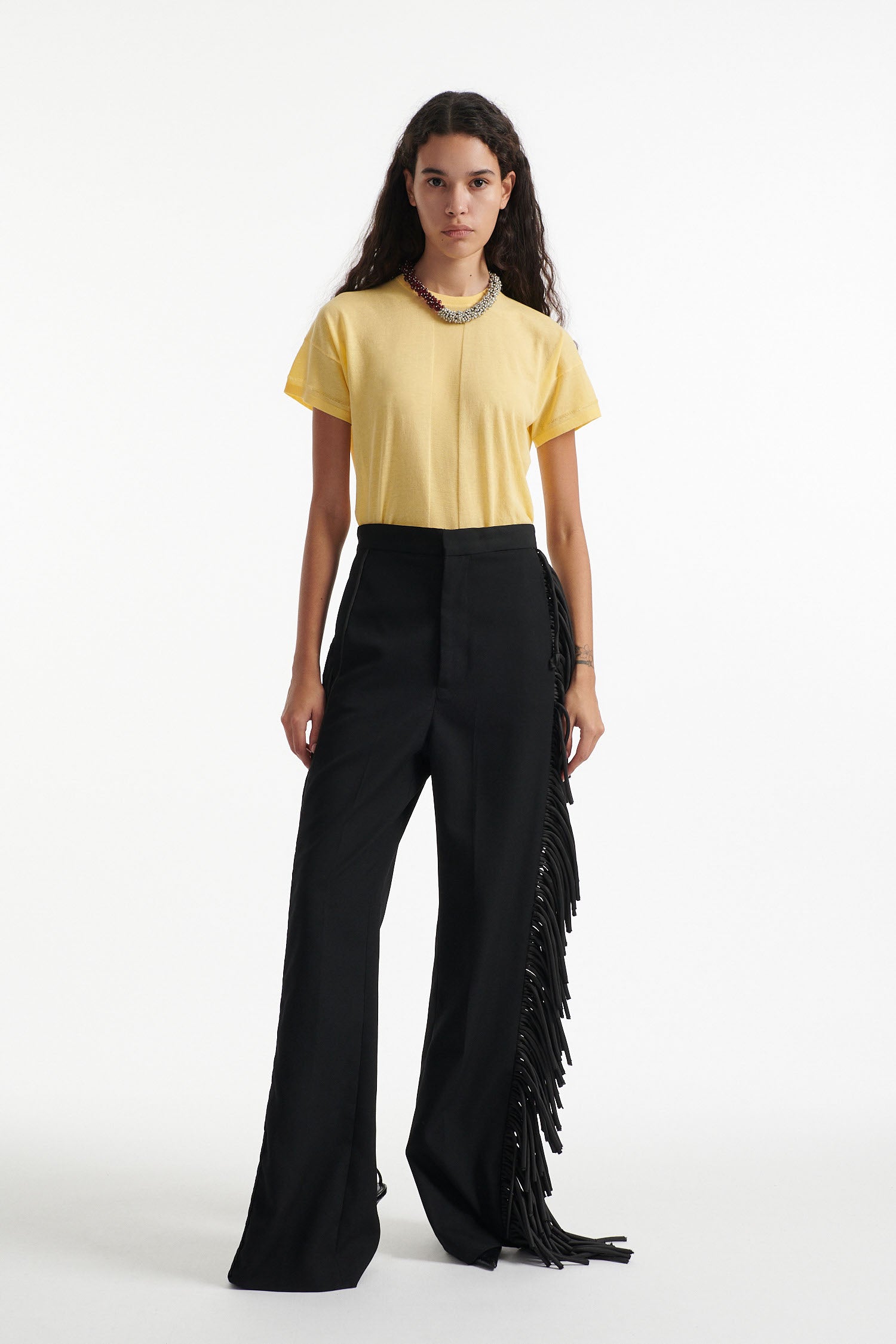 Female model wears long black fringe tuxedo pants with thin yellow cotton shirt and black high heels on white background. 