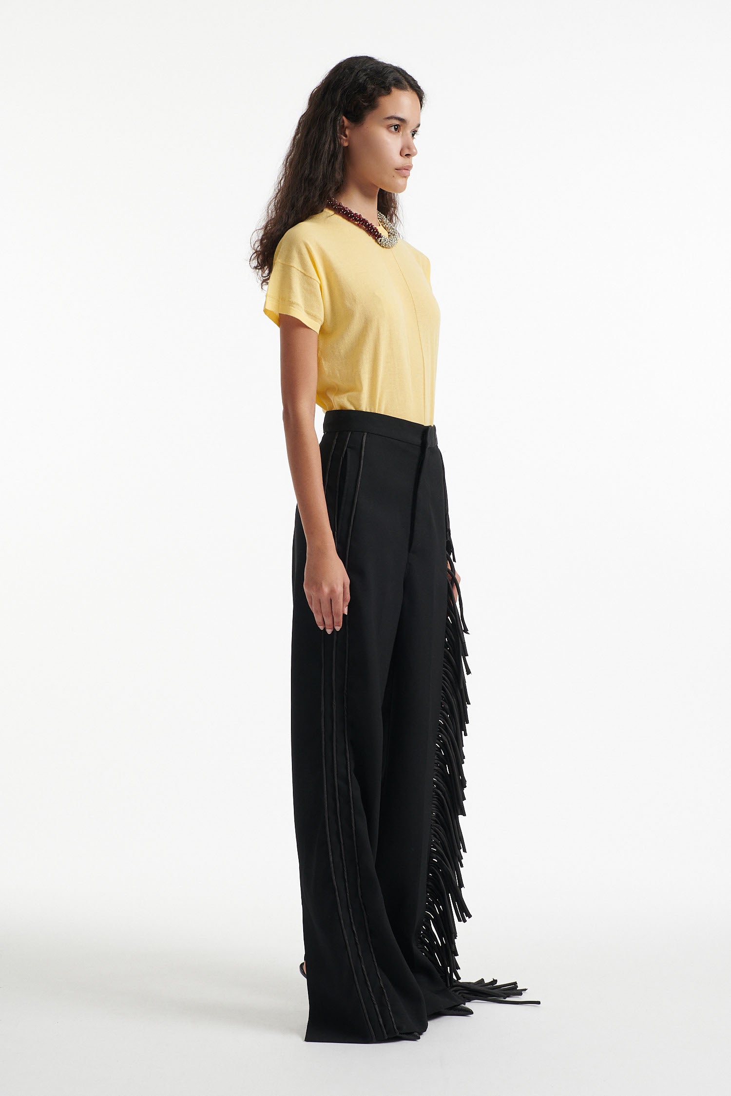 Female model wears long black fringe tuxedo pants with thin yellow cotton shirt and black high heels on white background. 