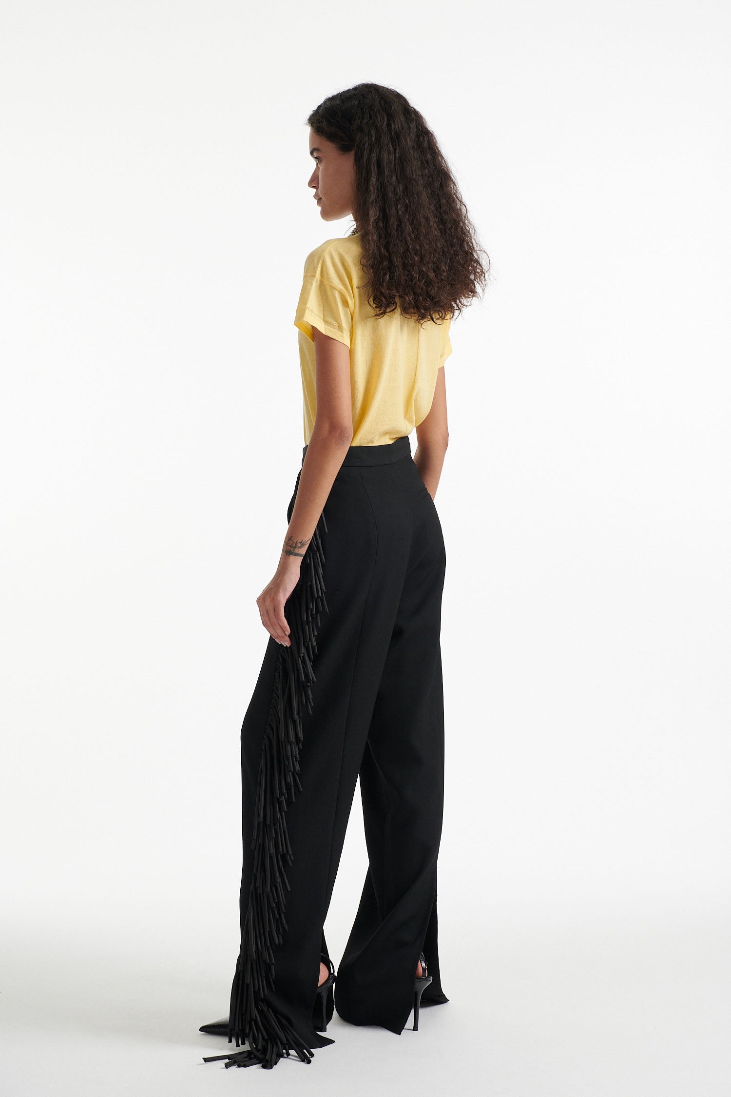 Female model wears long black fringe tuxedo pants with thin yellow cotton shirt and black high heels on white background. 