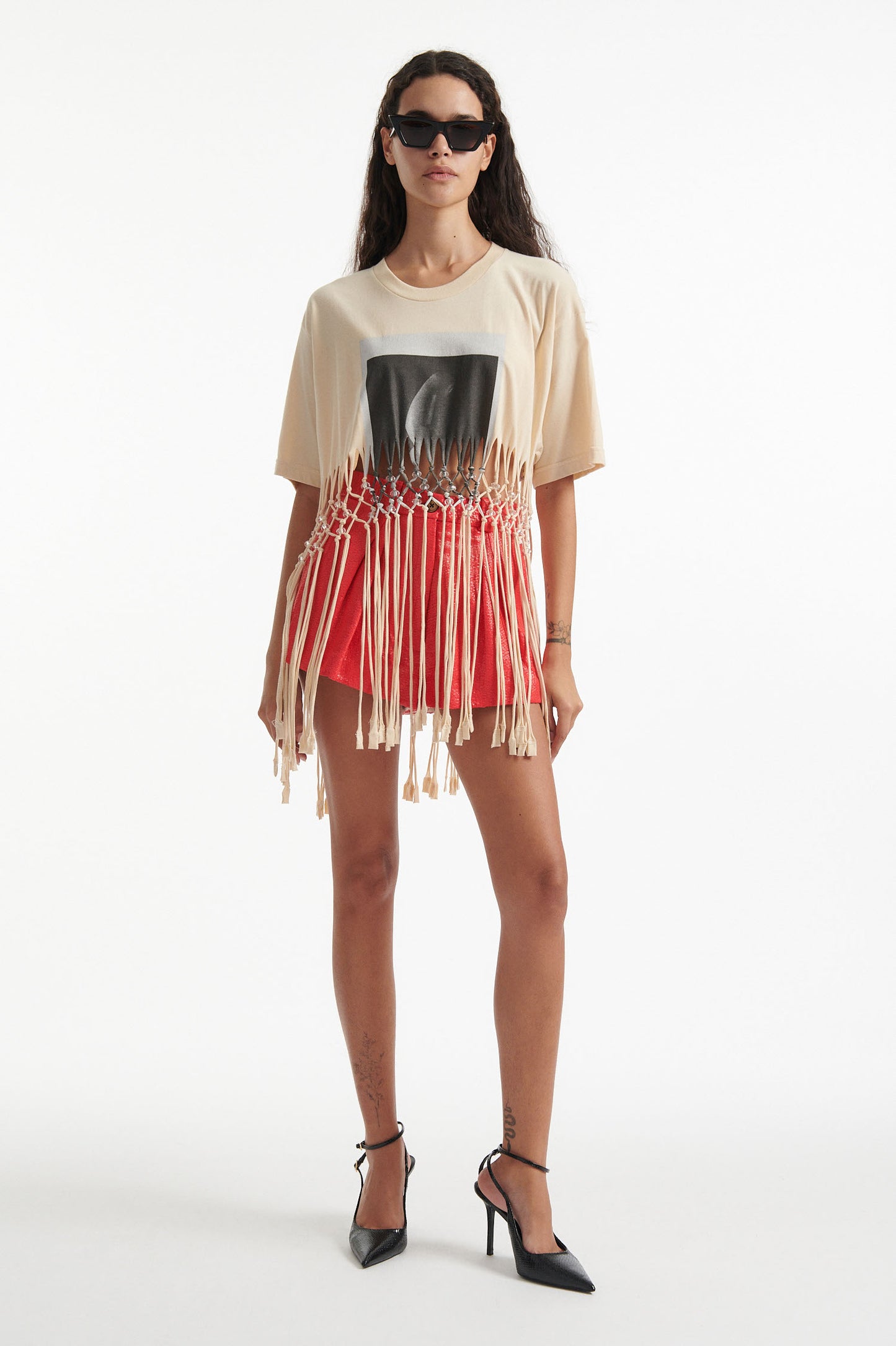 Female model wears deconstructed beige graphic tee with crystal embellishments and red shorts and black high heels on white background. 