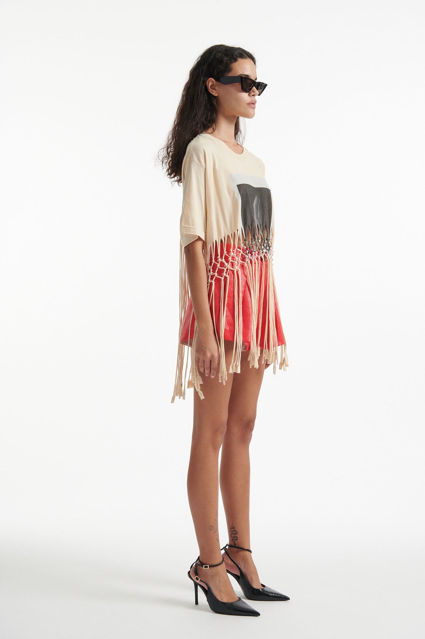 Female model wears deconstructed beige graphic tee with crystal embellishments and red shorts and black high heels on white background. 