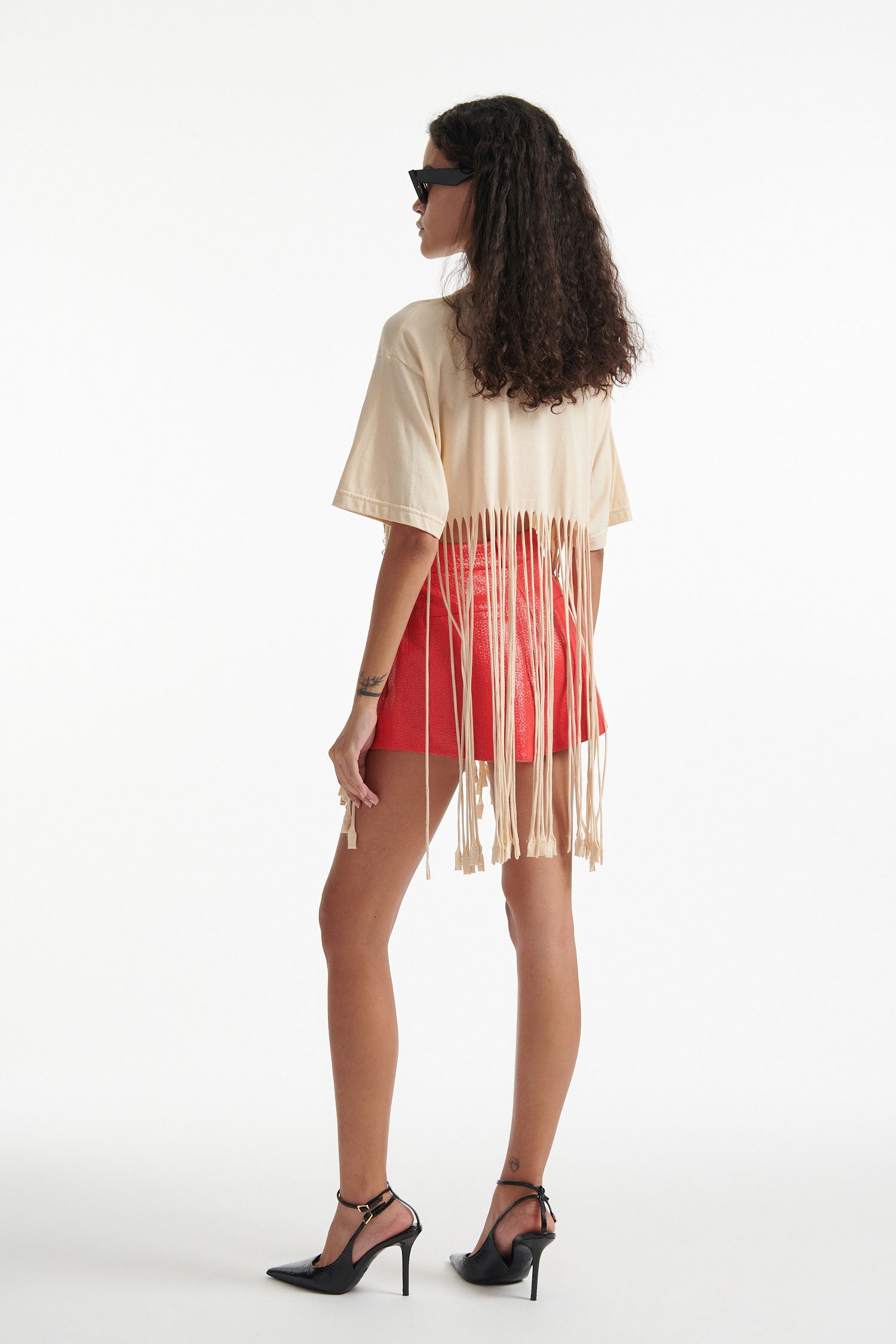 Female model wears deconstructed beige graphic tee with crystal embellishments and red shorts and black high heels on white background. 