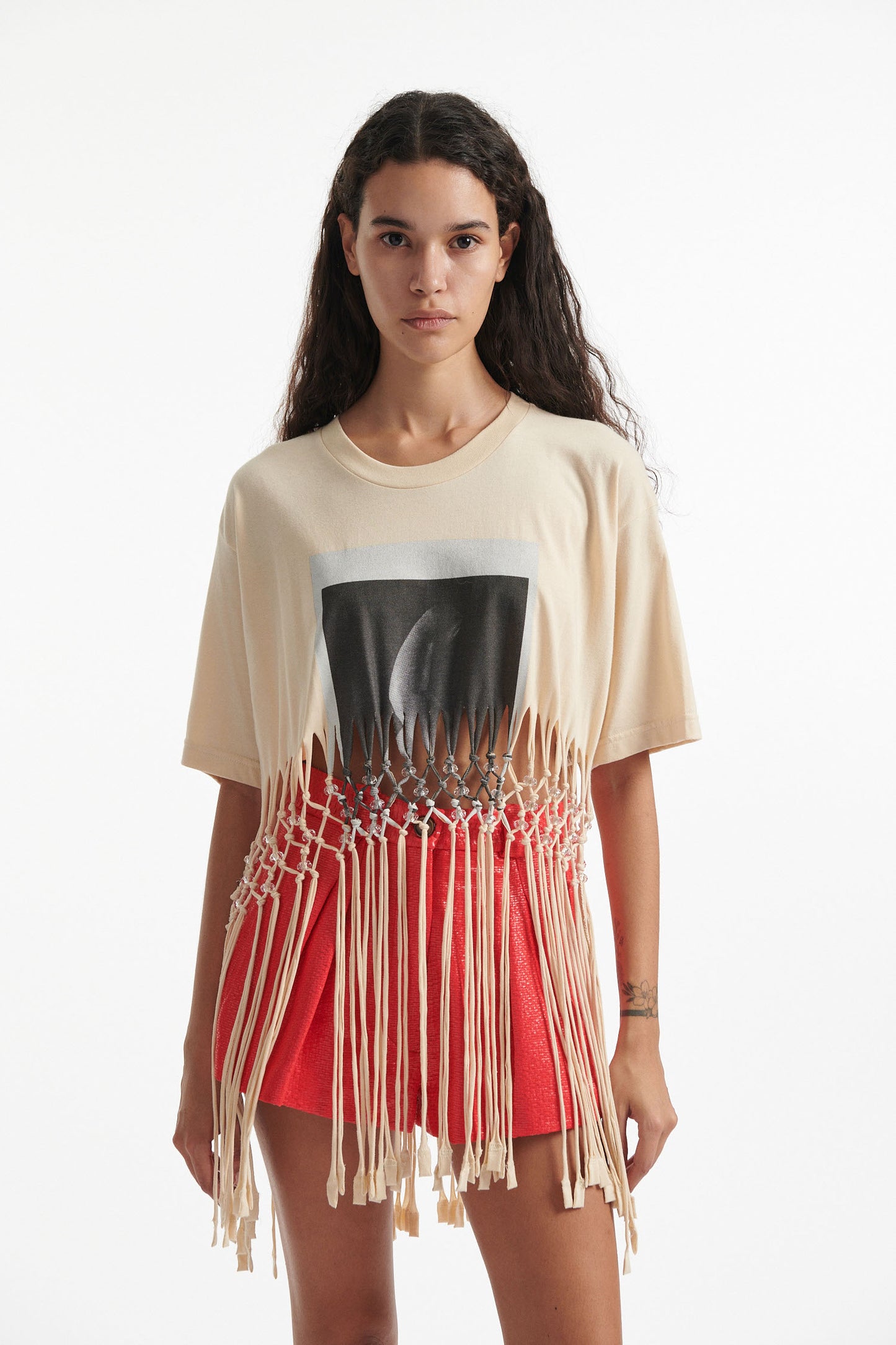 Female model wears deconstructed beige graphic tee with crystal embellishments and red shorts and black high heels on white background. 